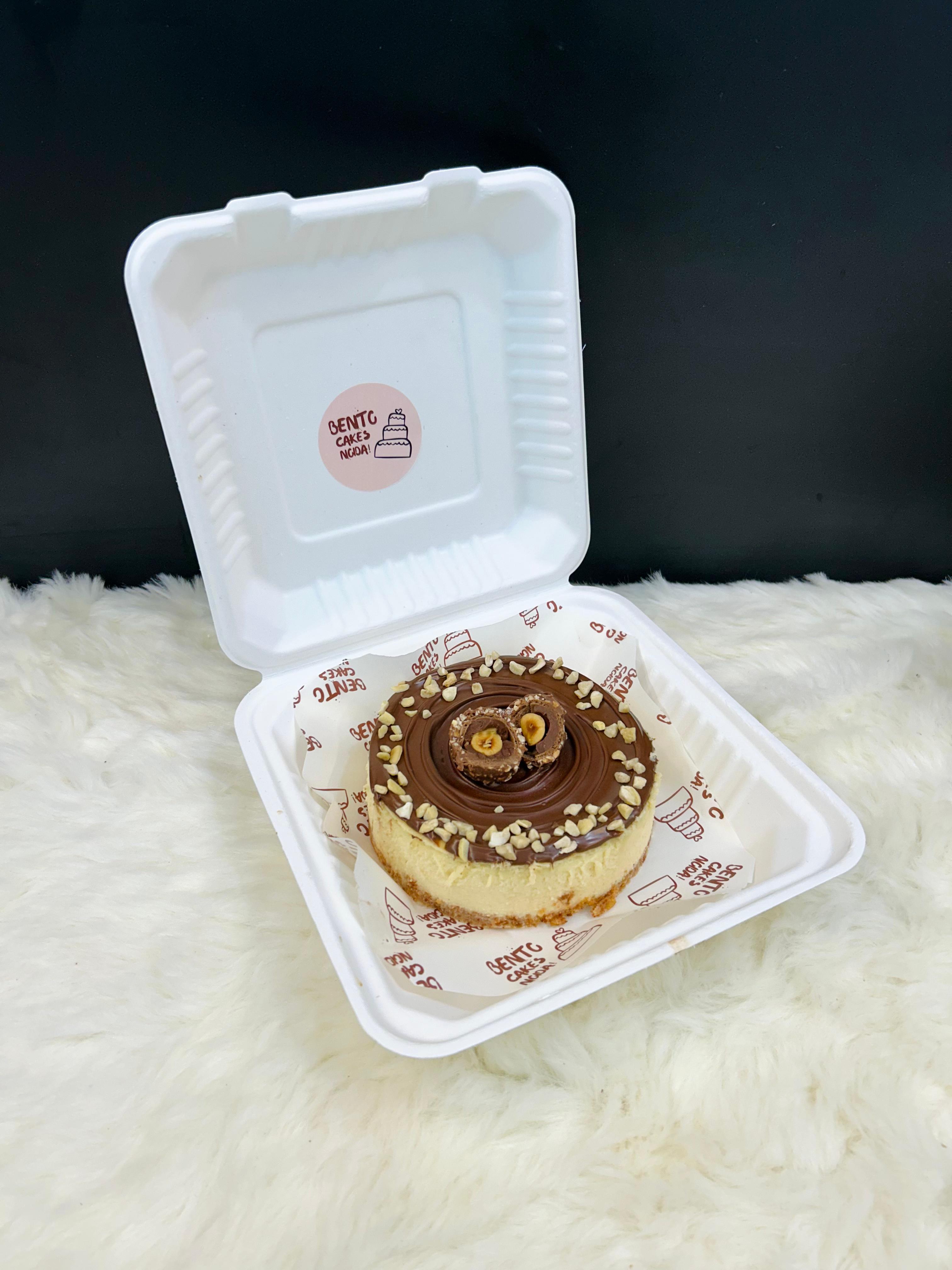 A luscious Nutella Cheesecake Bento in a white takeout box, topped with a glossy Nutella layer, Ferrero Rocher chocolates, and a sprinkle of chopped nuts, presented on a soft, white surface.