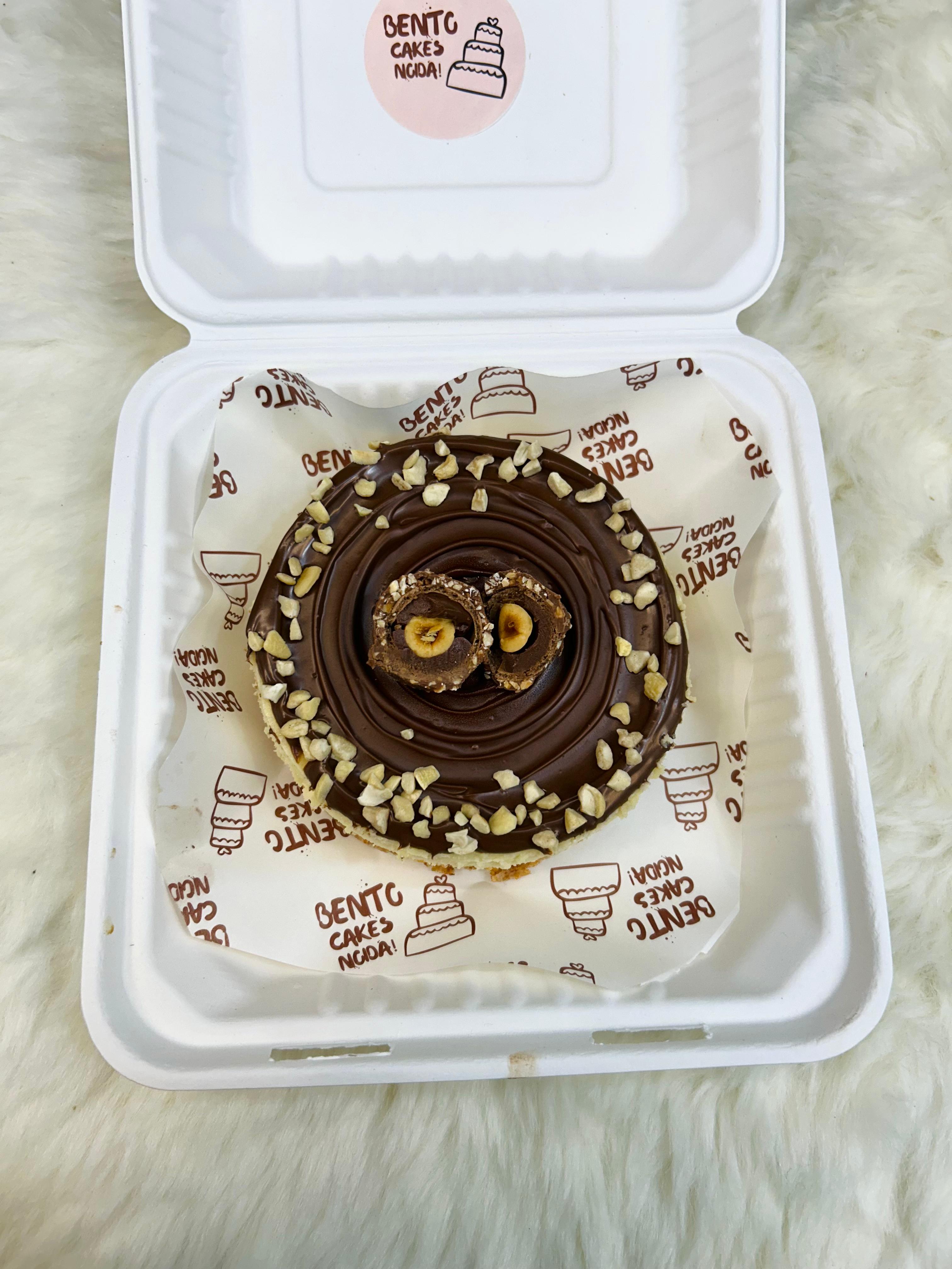 A luscious Nutella Cheesecake Bento in a white takeout box, topped with a glossy Nutella layer, Ferrero Rocher chocolates, and a sprinkle of chopped nuts, presented on a soft, white surface with front View.