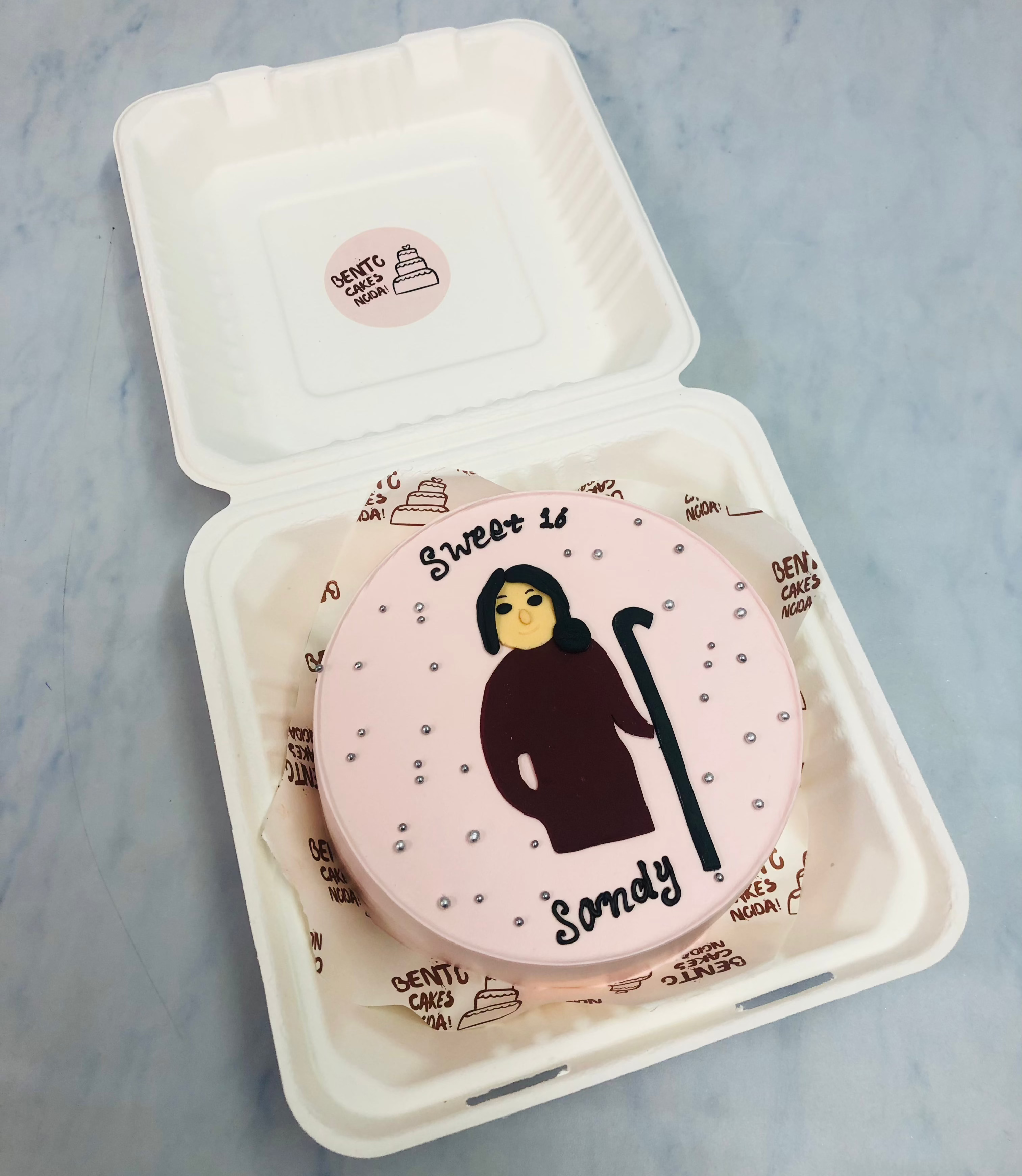 A pink round cake in a white box, decorated with a silhouette of a person holding a cane, and the text 'Sweet 16 Sandy'.