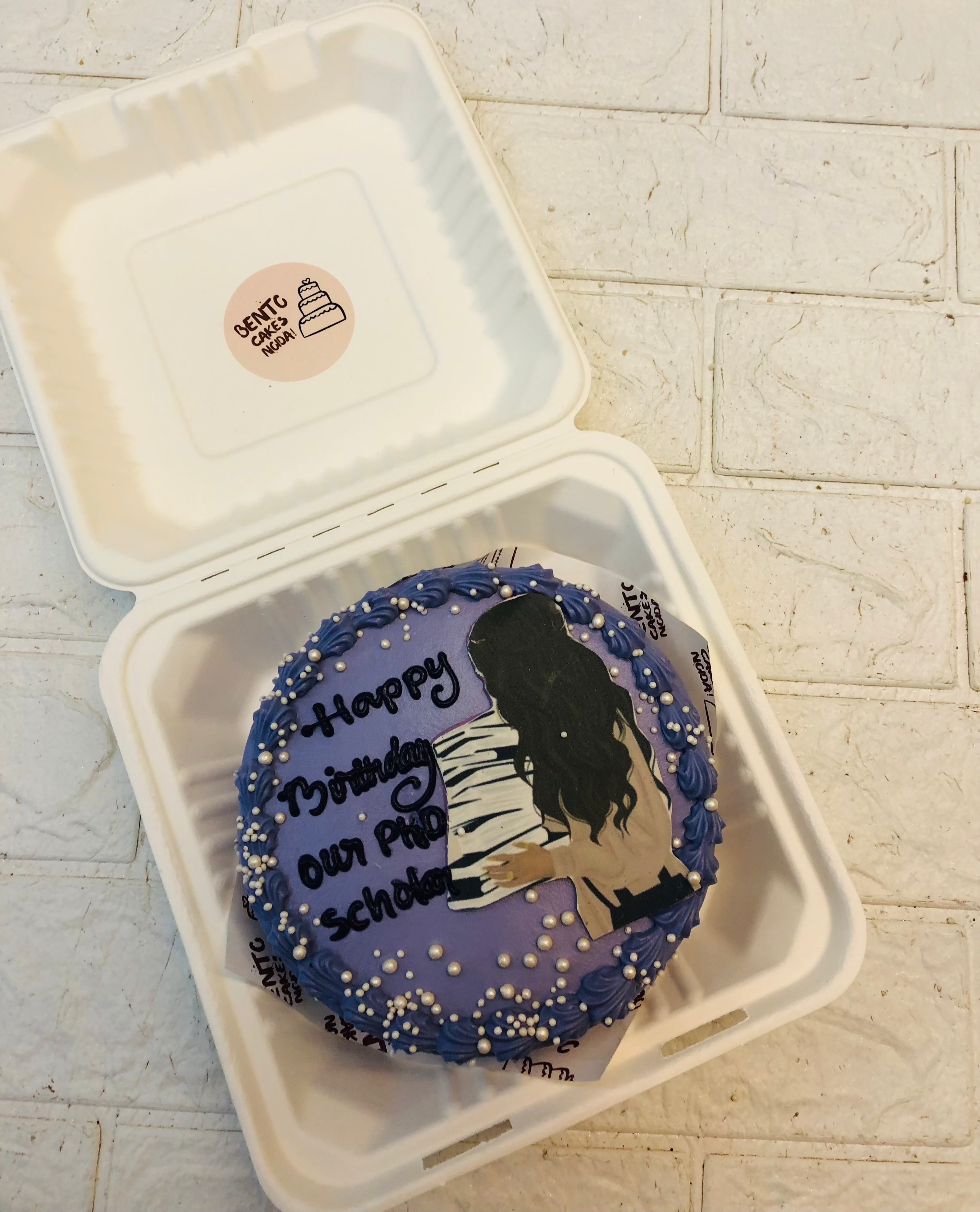 A beautifully decorated purple birthday cake featuring a detailed design of a girl playing the piano, placed inside a white bento box. The cake is adorned with white and blue icing and small white pearls, with the text 'Happy Birthday Our Phd Scholar' elegantly displayed on top.