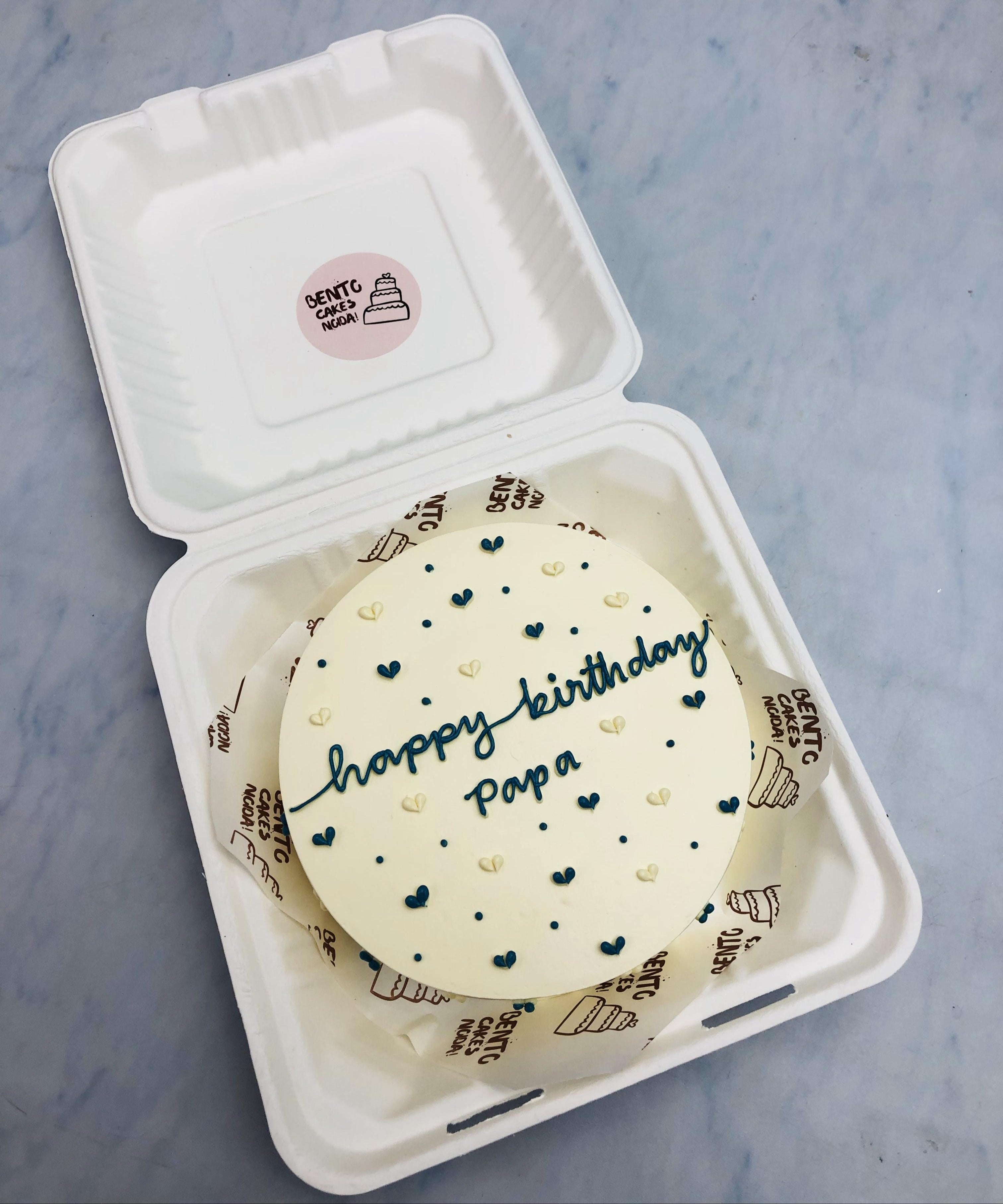 Little blue and white hearts designed on an offwhite bento cake with a text "happy birthday papa" written with blue icing.