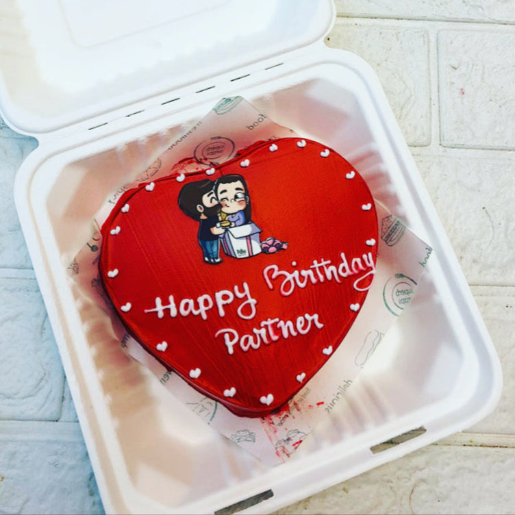 Heart-shaped birthday cake with a cartoon couple illustration and the text "Happy Birthday Partner" in a white bento box.