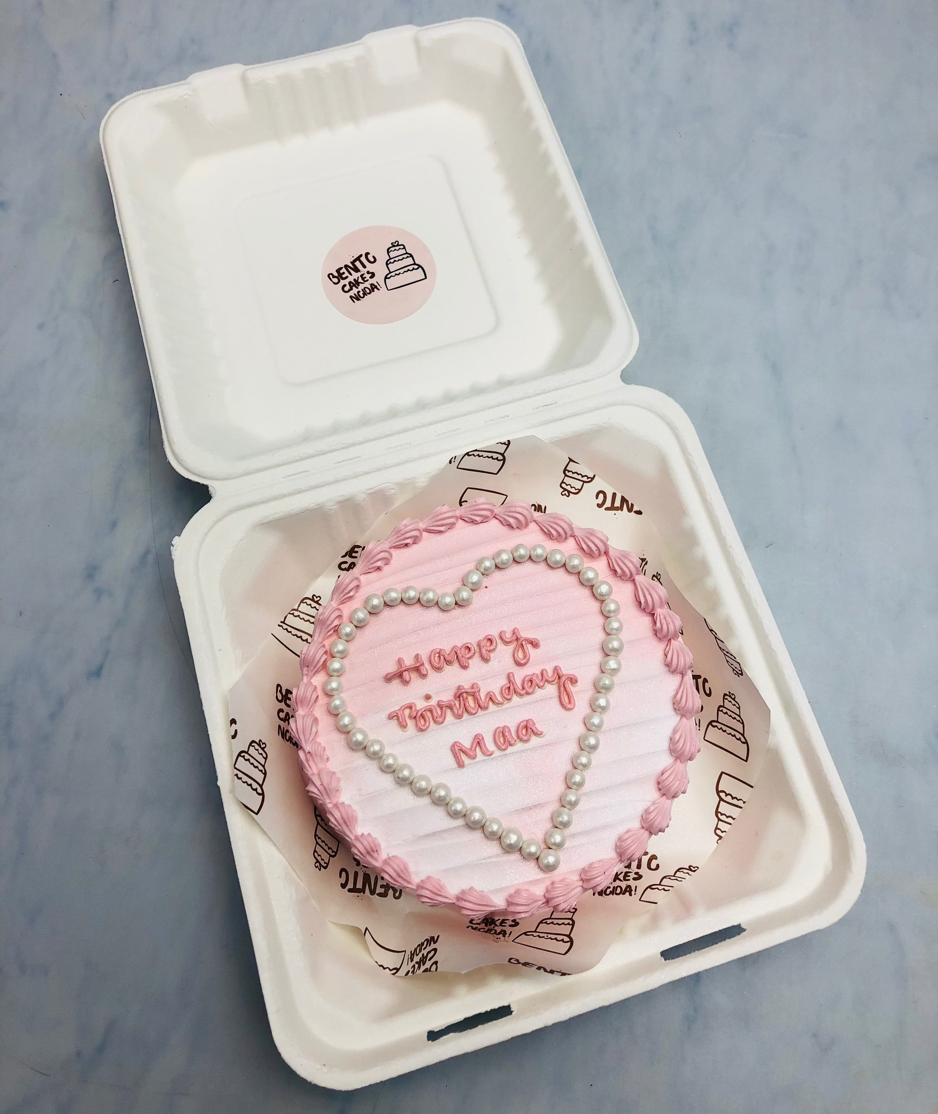 A pink heart-shaped birthday cake with the message 'Happy Birthday Maa' written in pink icing, decorated with pearl-like beads around the heart, placed inside a bento box with branded paper lining.