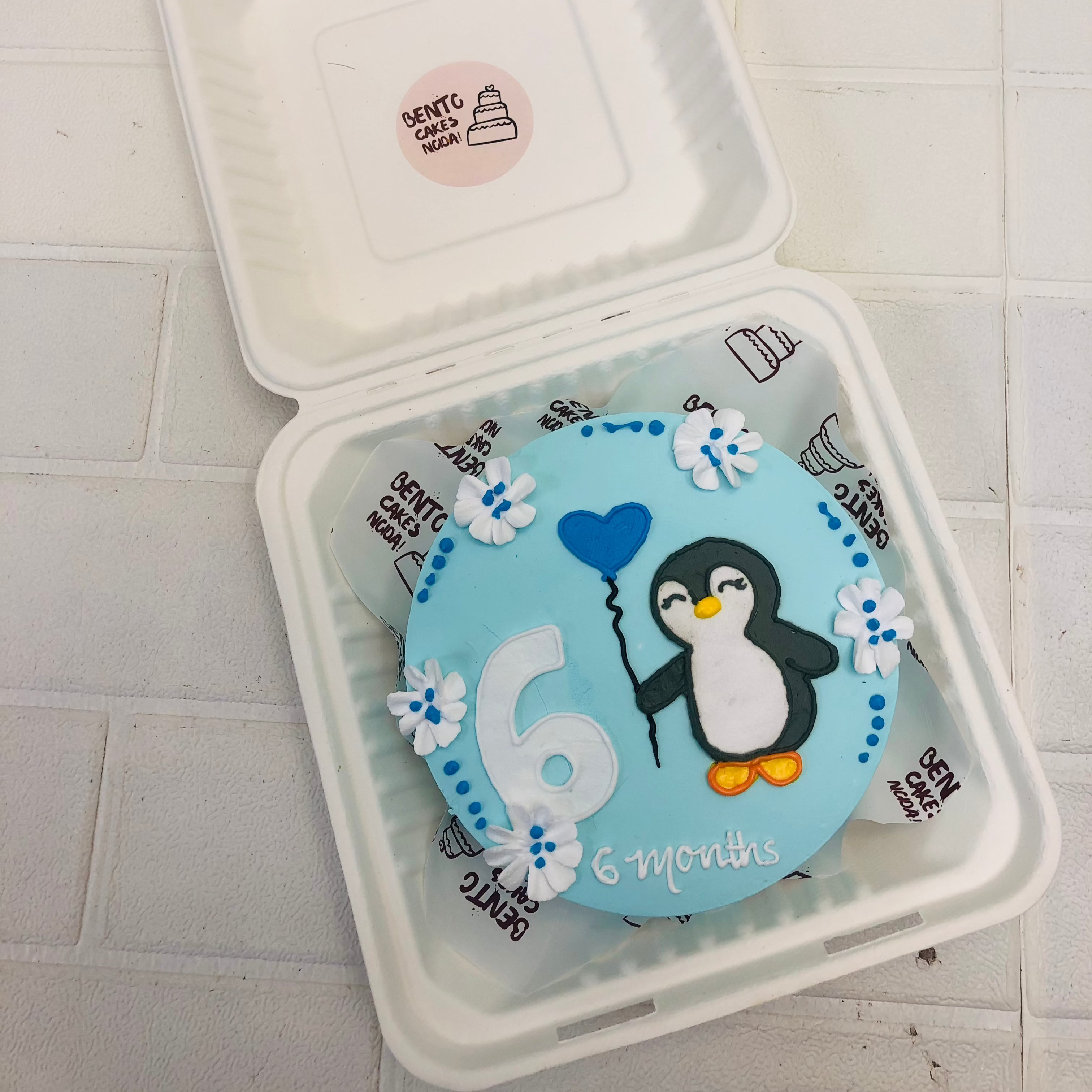 A light blue bento cake with cute panguine design with balloon in hand.