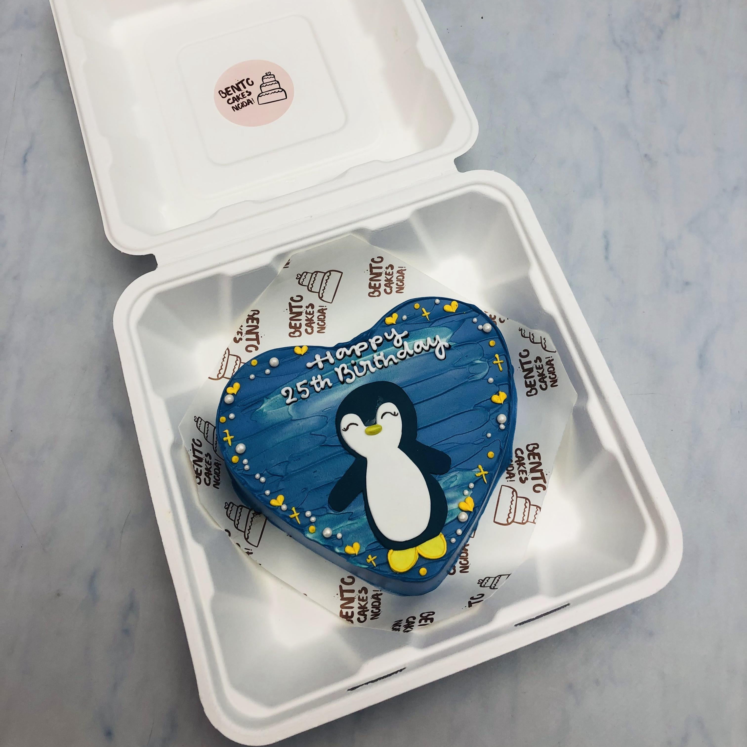 An aesthetic blue heart shape bento cake with a cute panguin design on it.
