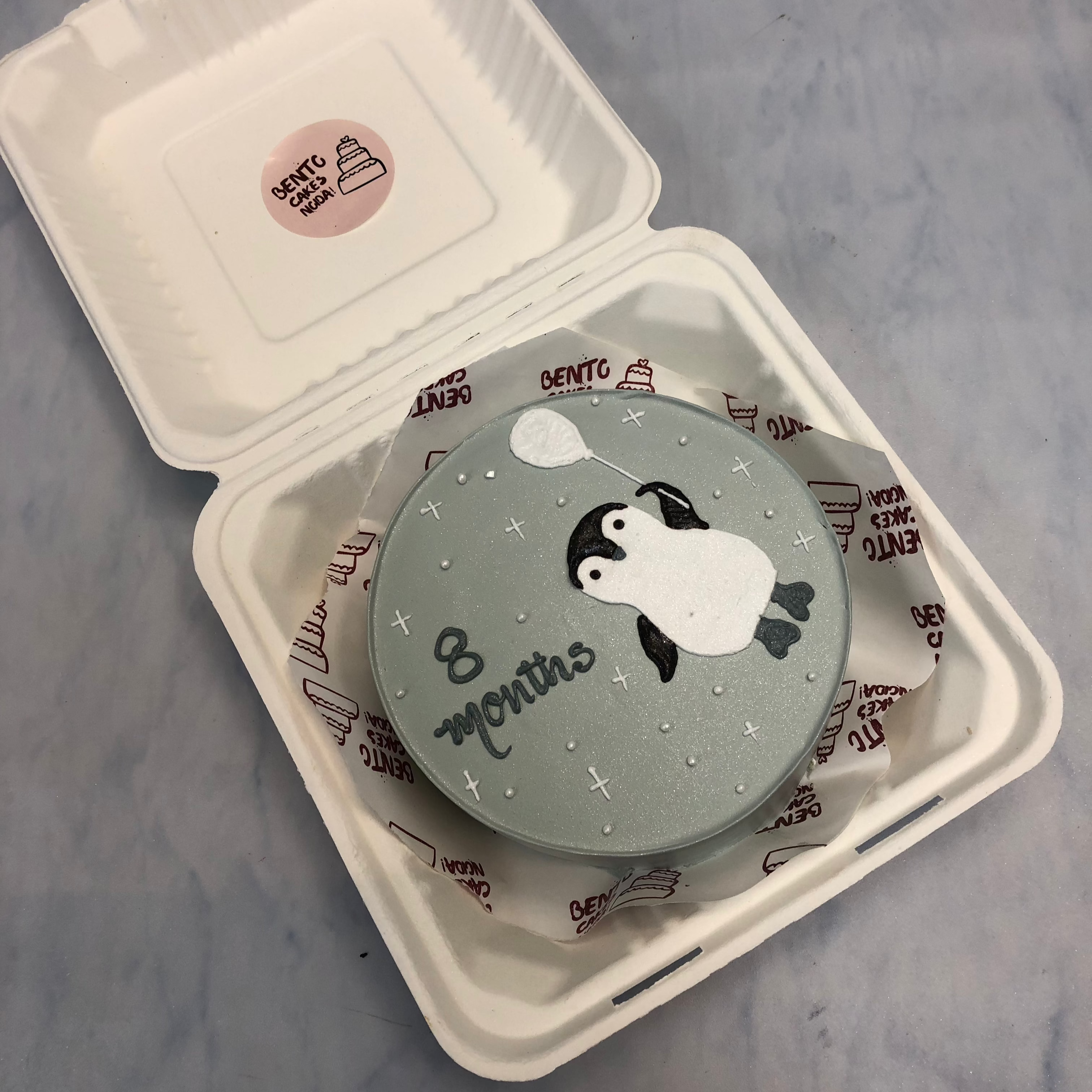 A grey bento cake with a cute little panguine taken balloon and white stars filled all over the cake.