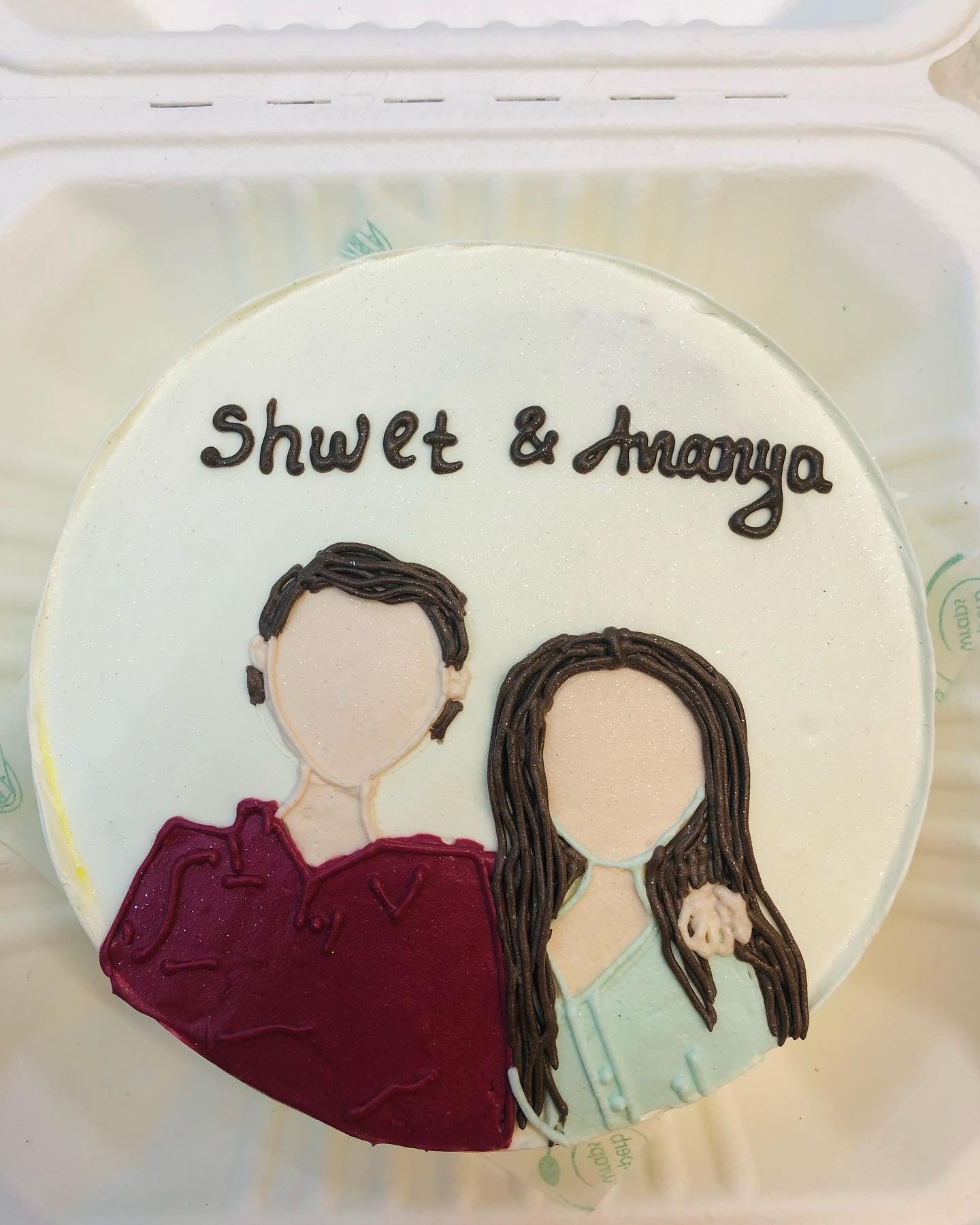 A round cake with a white frosting base featuring a minimalist illustration of two people, one in a maroon top and the other in a light blue top, with the names "Shwet & Ananya" written in chocolate icing at the top.