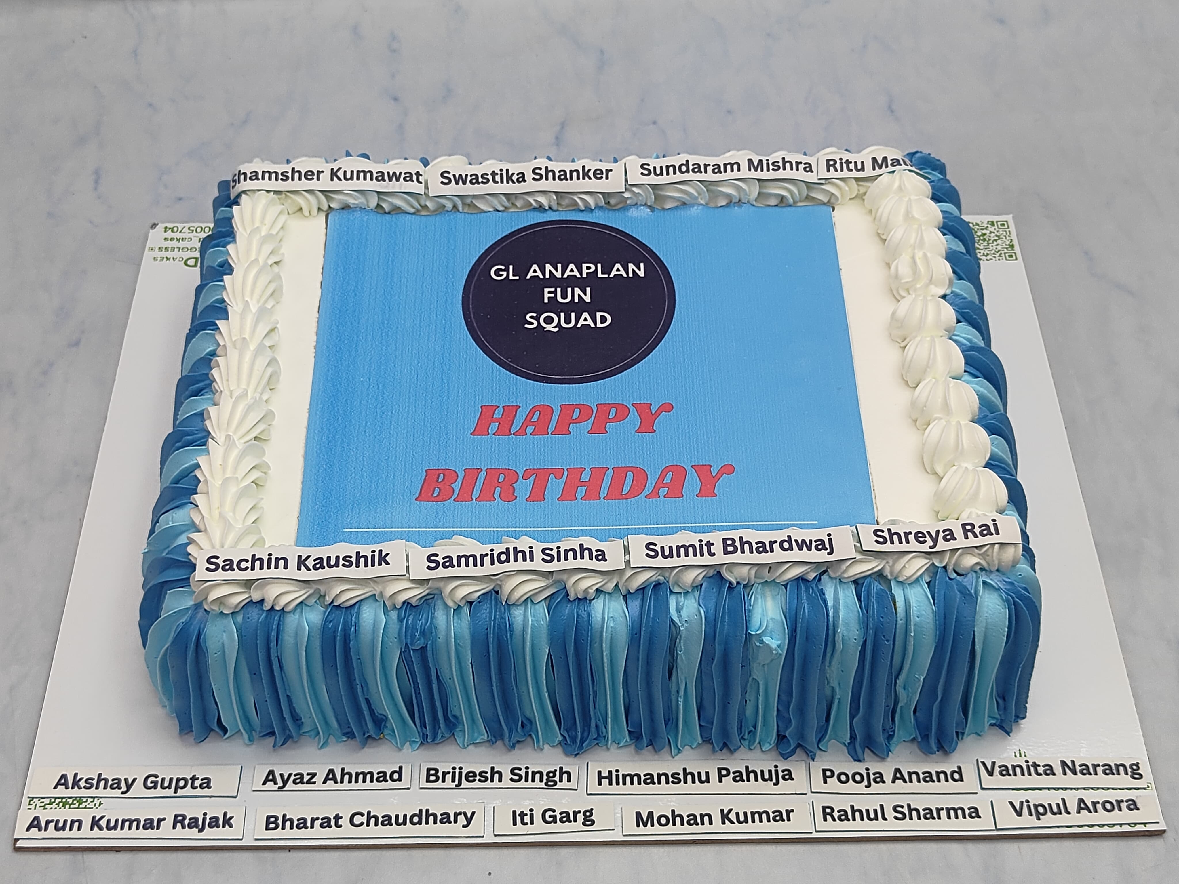 A rectangular birthday cake with blue and white icing, featuring the text "GL ANAPLAN FUN SQUAD" and "HAPPY BIRTHDAY" on top. The cake is decorated with the names of team members around the edges.