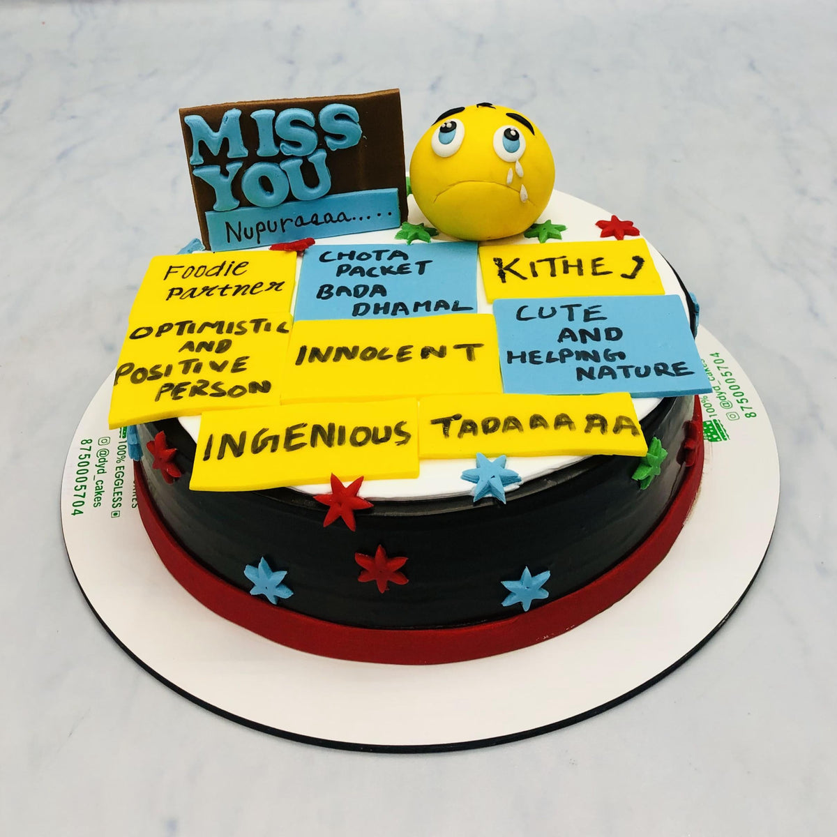 A round cake with a black and red base, decorated with colorful stars and various yellow and blue sticky notes with handwritten messages. The cake features a crying emoji face and a sign that reads "MISS YOU Nupuraaaa...."
