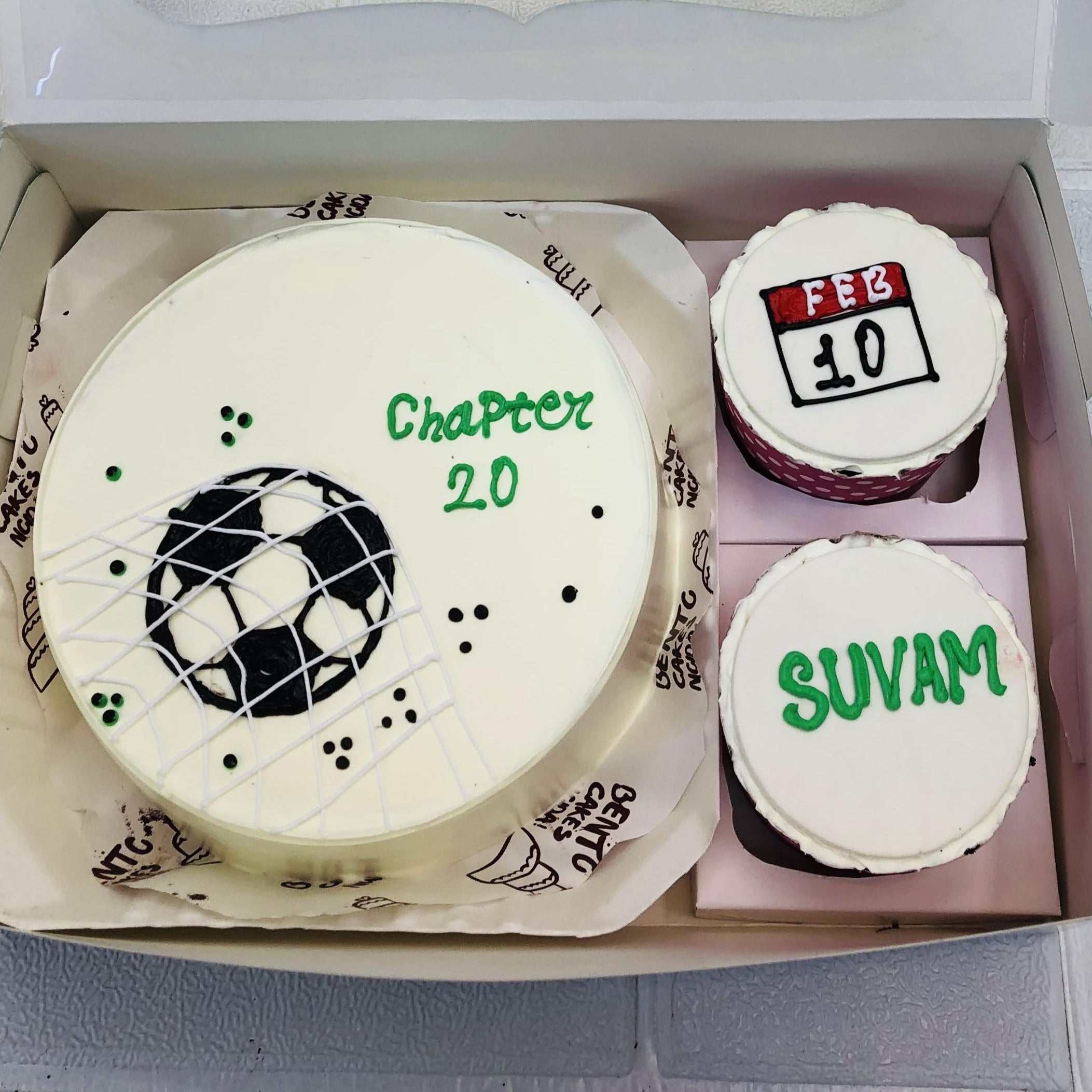 A set of cakes in a box, including a soccer-themed large round cake with the text "Chapter 20," and two smaller round cakes, one featuring the date "Feb 10" and the other the name "Suvam," all intricately designed and decorated.