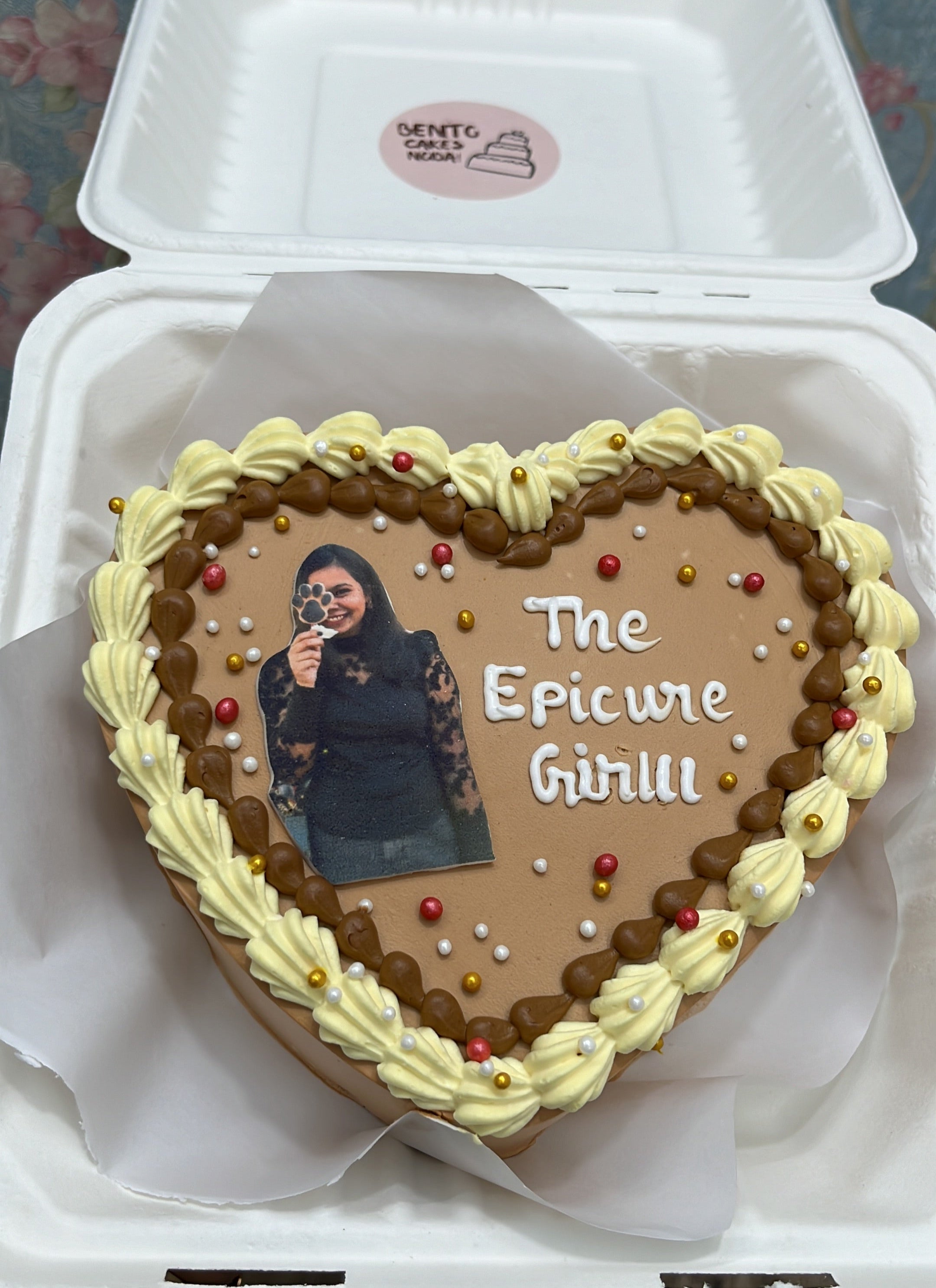 A heart-shaped cake with personalized decoration, featuring a photo of a person and the text "The Epicure Girlll." The cake is adorned with cream and chocolate frosting, along with colorful edible pearls.