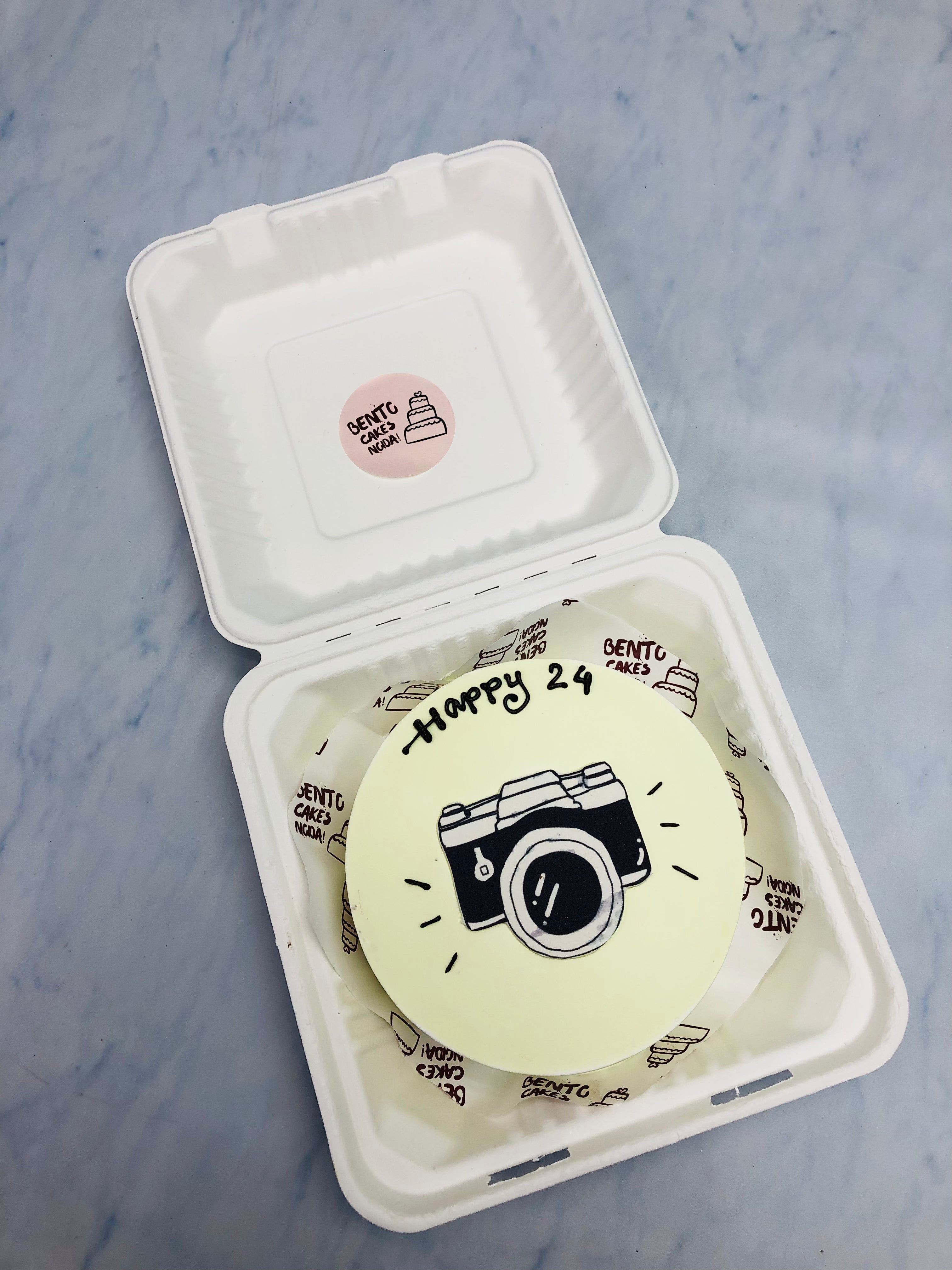 A light yellow bento cake is designed for 24th birthday with camera print.