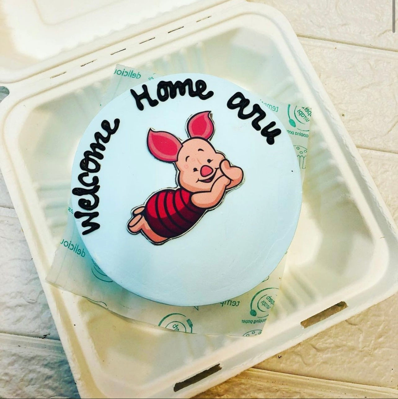 A light blue cake designed with a sleepy carton pig sticker on top and a text "Welcome Home aru" written with black icing above the sticker and is placed inside a bento box.