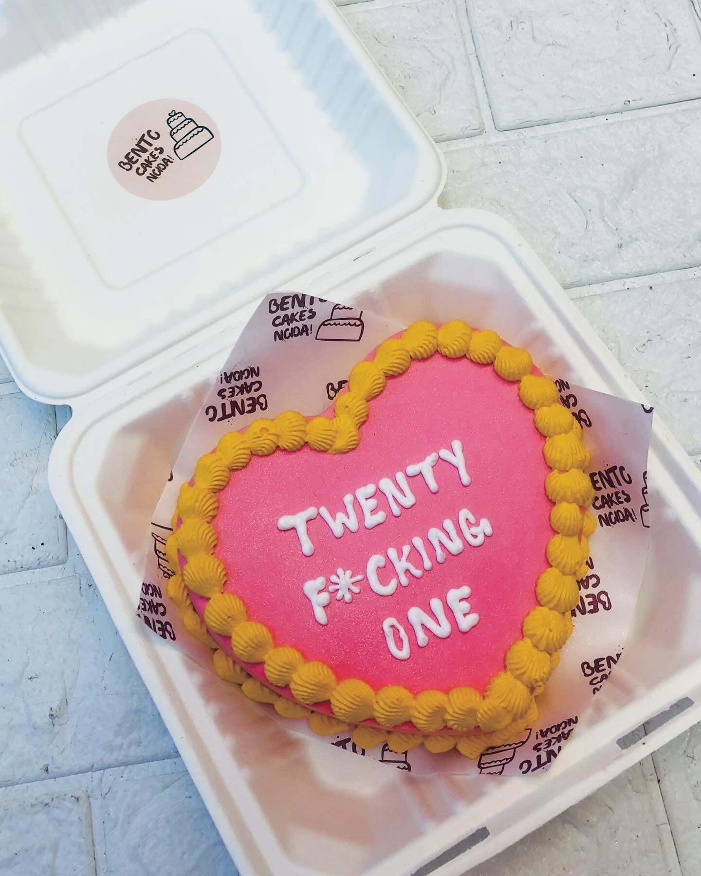 A shiney pink heart shape cake with yellow boundry design on it's top and bottom and a text written with white icing on it.