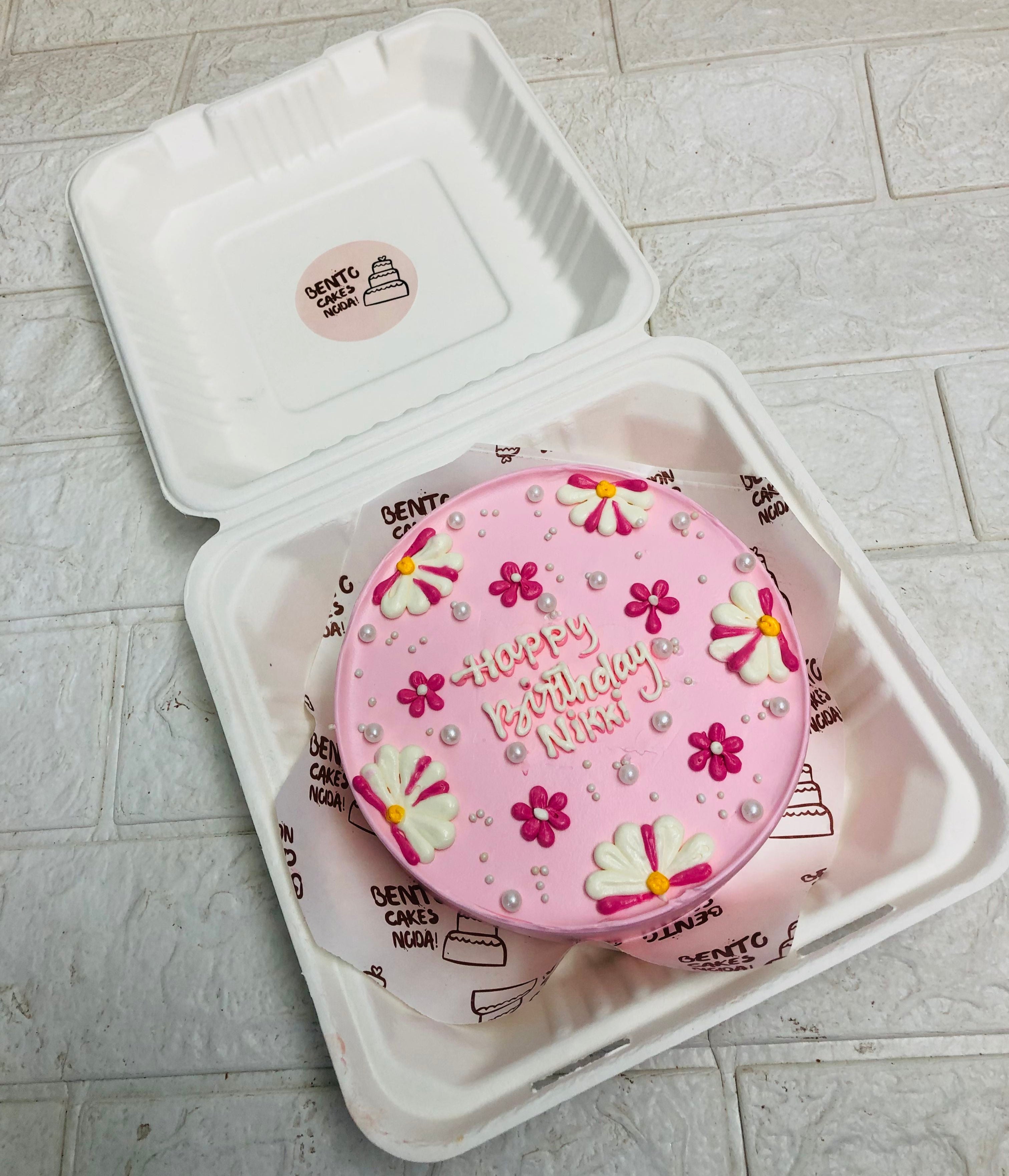 A beautifully crafted pink bento cake featuring floral decorations and the message 'Happy Birthday Nikki.