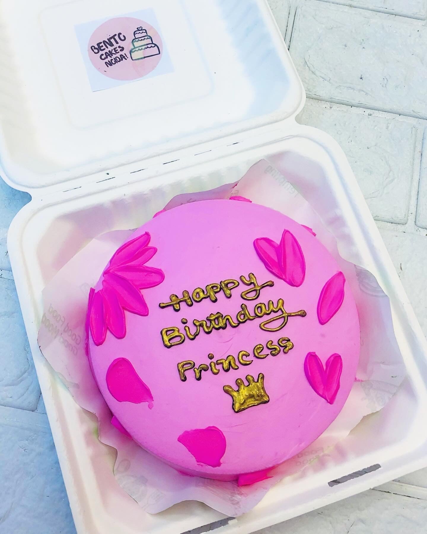A pink birthday cake with the words "Happy Birthday Princess" written in gold icing, surrounded by pink heart and flower decorations, placed inside a white box. The cake is topped with a gold crown decoration.