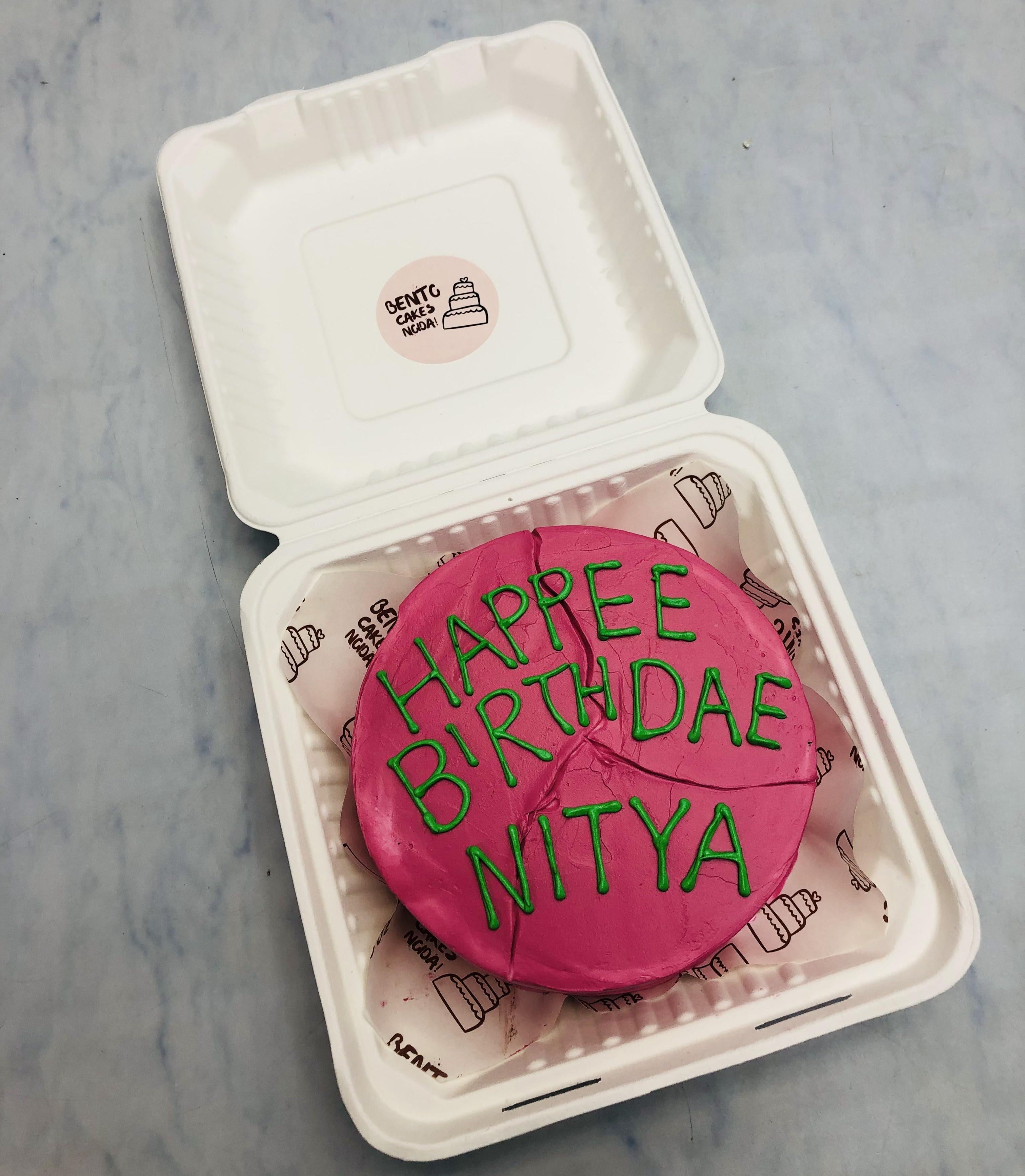 A pastel pink birthday cake with whimsical green lettering spelling 'HAPPEE BIRTHDAE NITYA,' placed inside a white takeout box. 