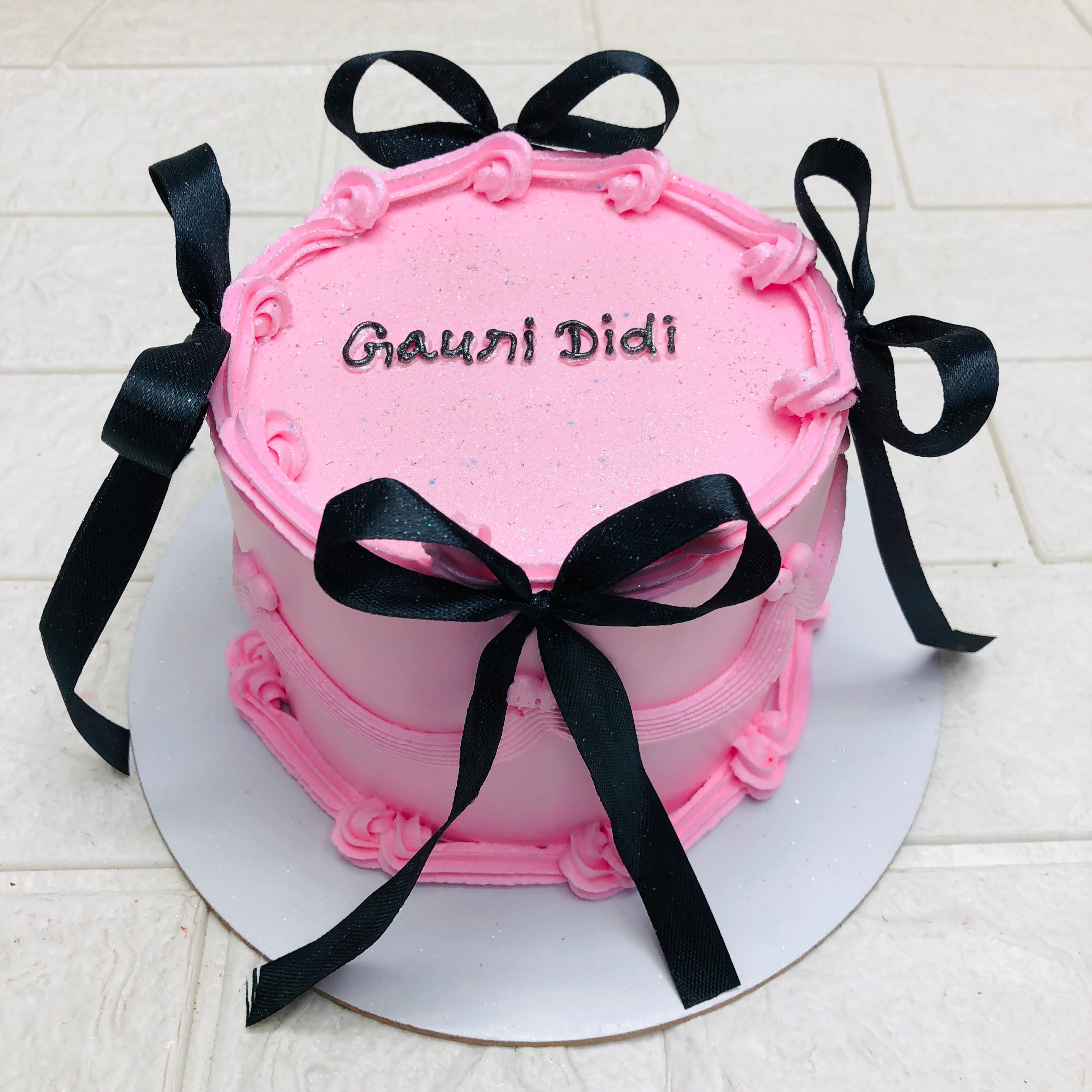 A round pink cake with black ribbon bows and the text "Gauri Didi" written on top in black icing.