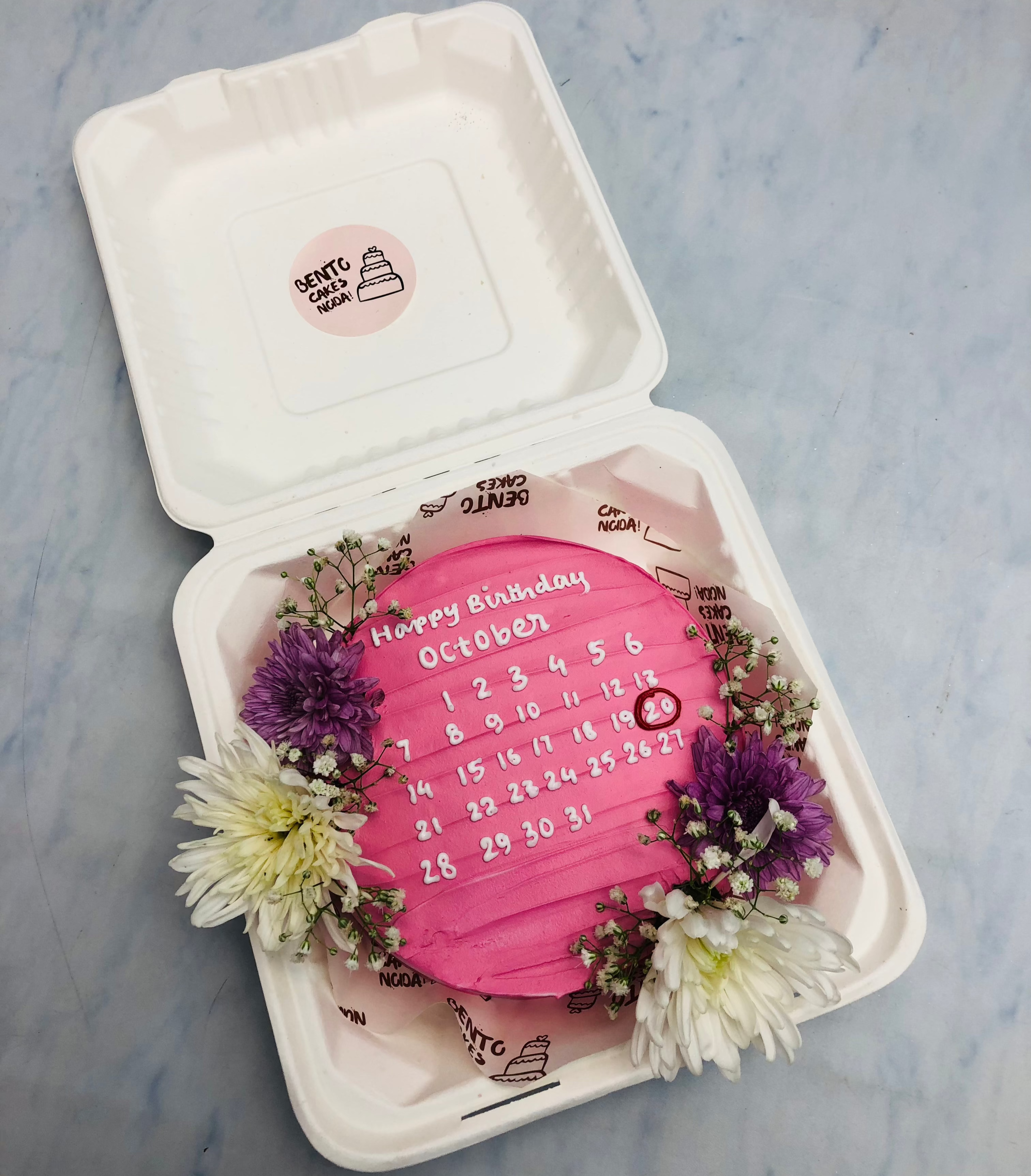 A pink bento cake with calendar design and decorated with fresh flowers.