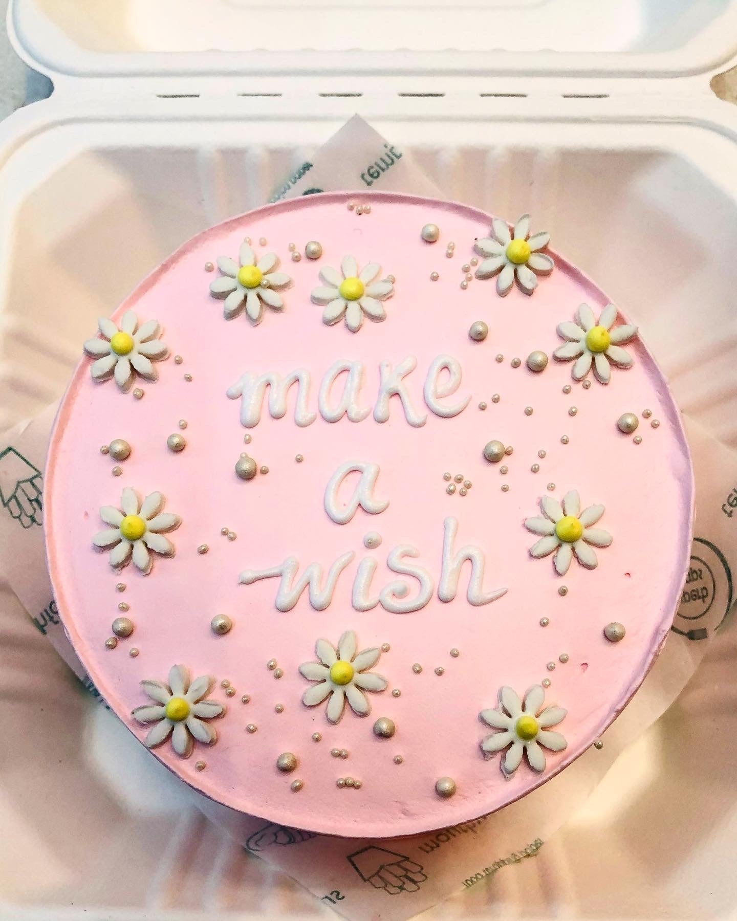 A round pink cake with the words 'make a wish' written in white icing, decorated with white and yellow daisy flowers and silver pearls.