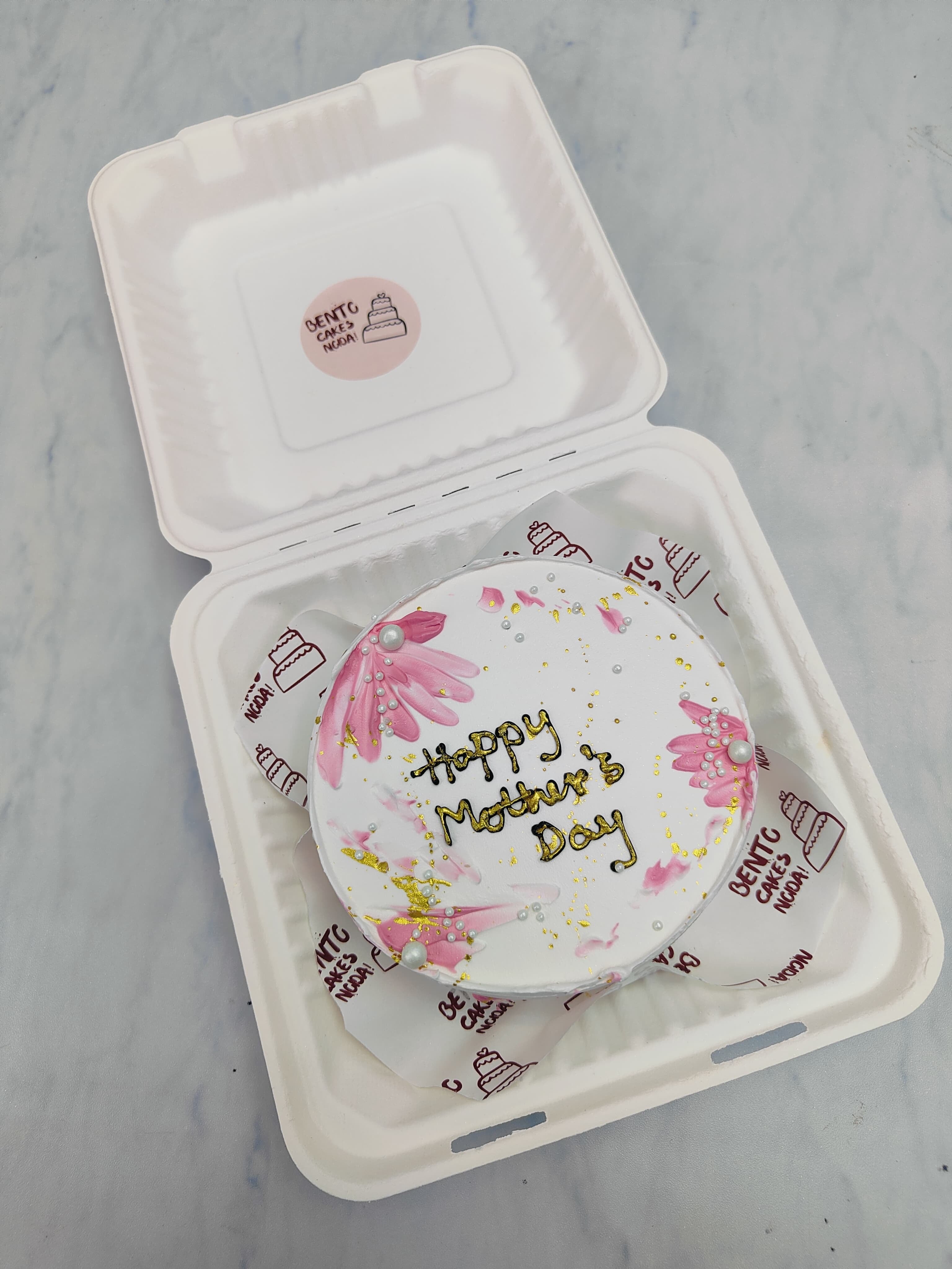 A white bento cake having creamy floral shades with golden text on it for mothers day occasion.