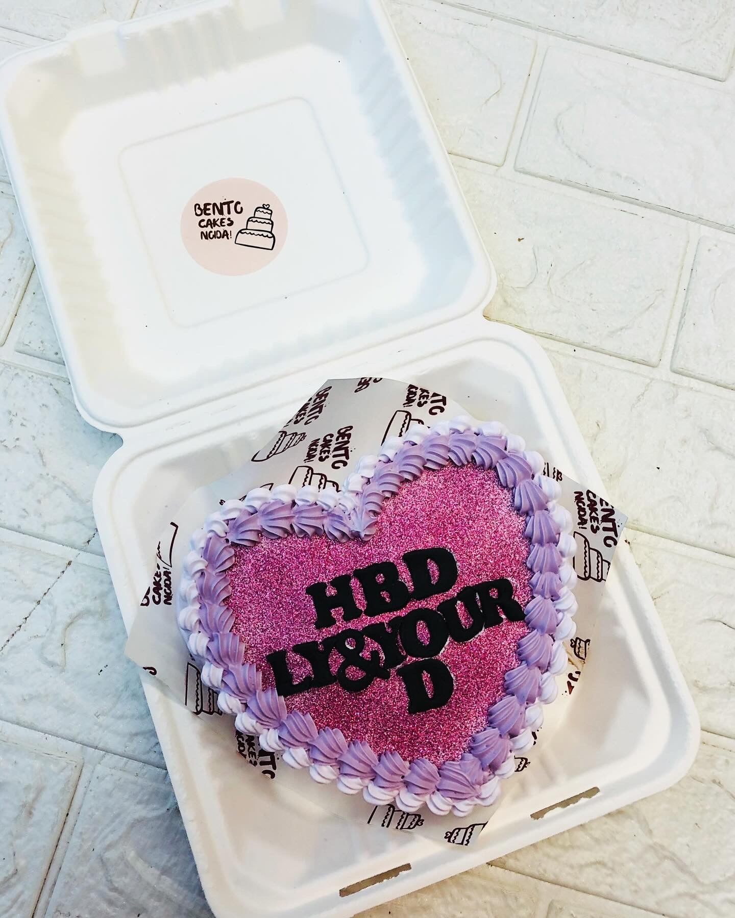 A purple glitter heart shape cake with purple and white designs on boundary and a text written with black fondant.