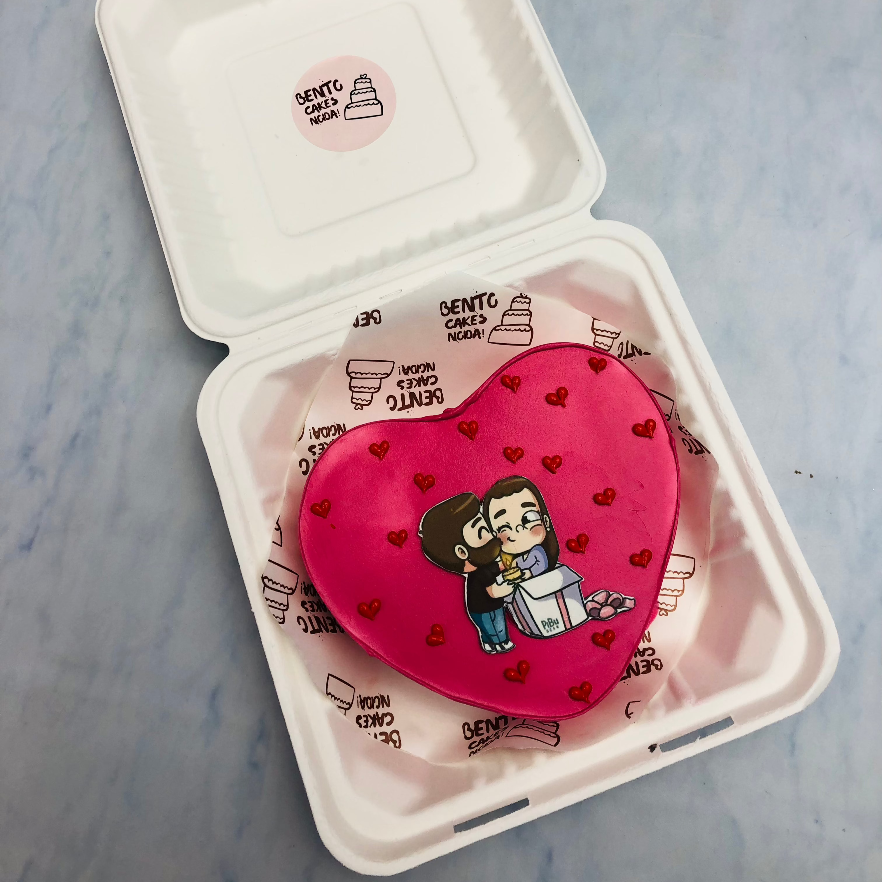 A dark pink heart shape cake with a kissing couple print, with red hearts.