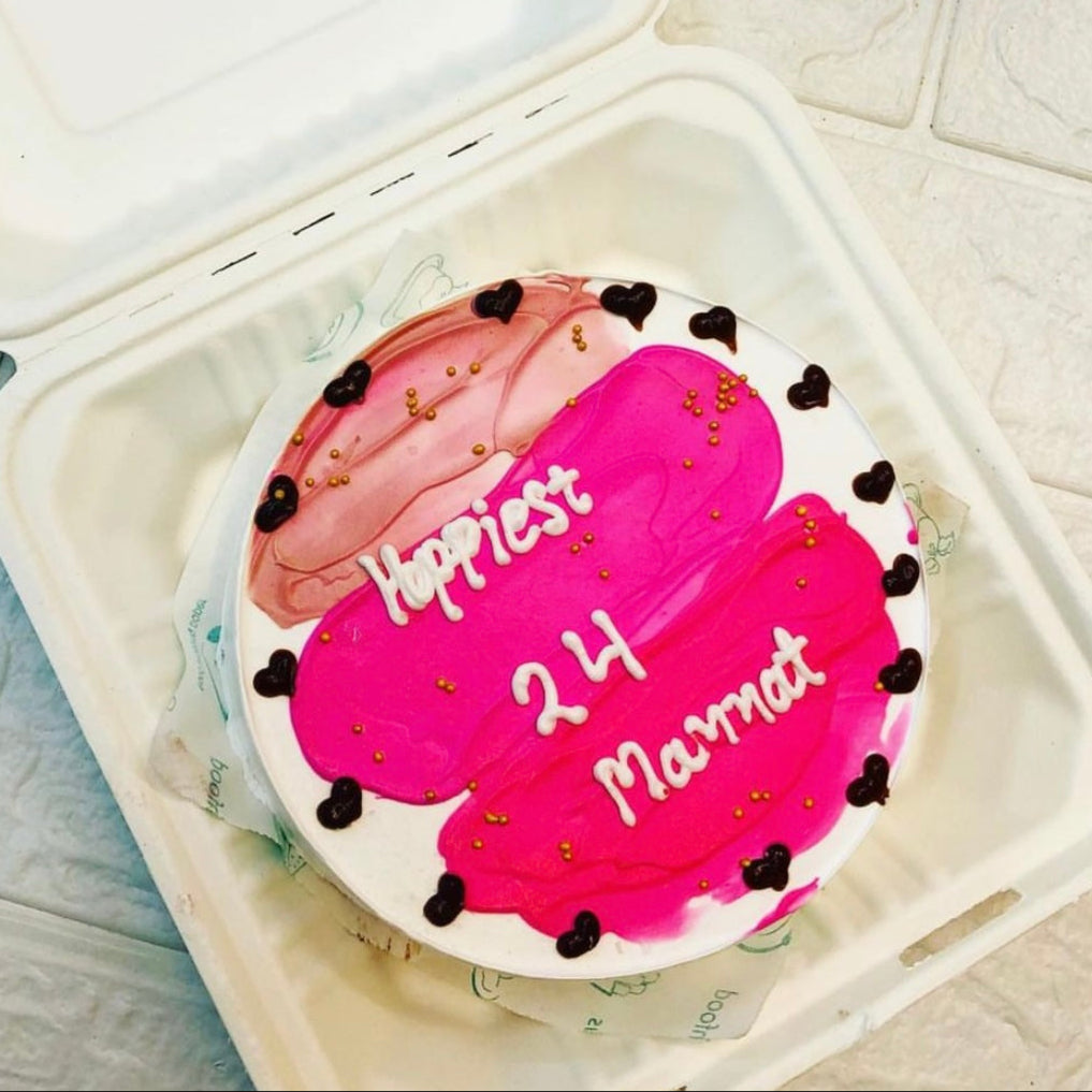 A round cake with pink and red frosting, decorated with small black heart shapes and golden sprinkles, placed inside a white bento box. The cake has the text "Happiest 24 Mannat" written in white icing.