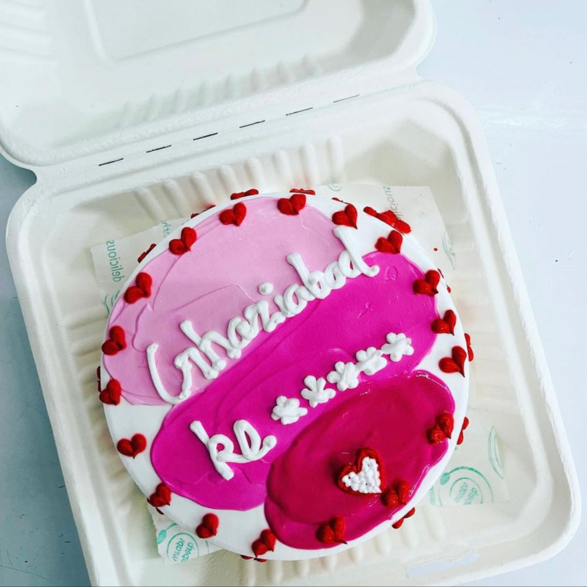 A round cake with pink and white frosting, decorated with small red hearts and white text that reads "Ghaziabad ke"