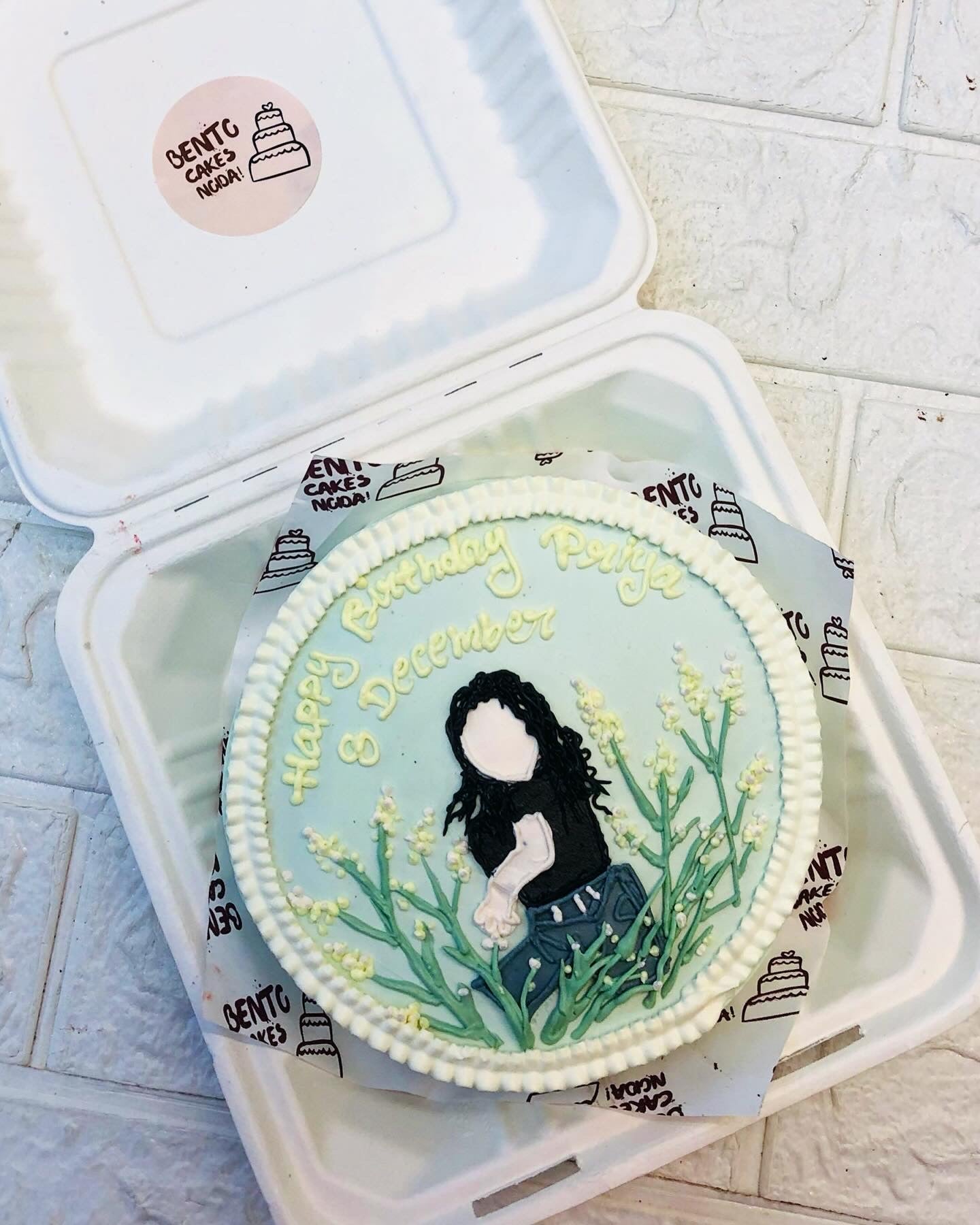 A Girl Theme Bento Cake, featuring a floral design and a custom message that reads "Happy Birthday Pooja 8 December". The cake is decorated with an illustration of a person with long black hair wearing a black shirt and jeans, surrounded by yellow flowers and green leaves.
