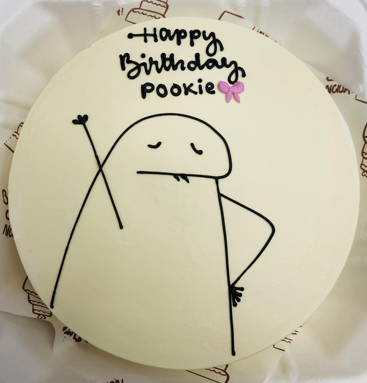 A round birthday cake with a minimalist design featuring a simple black line drawing of a character with closed eyes and a small tongue sticking out. The cake has the text "Happy Birthday pookie" written on top, with a small pink bow decoration next to the word "pookie."