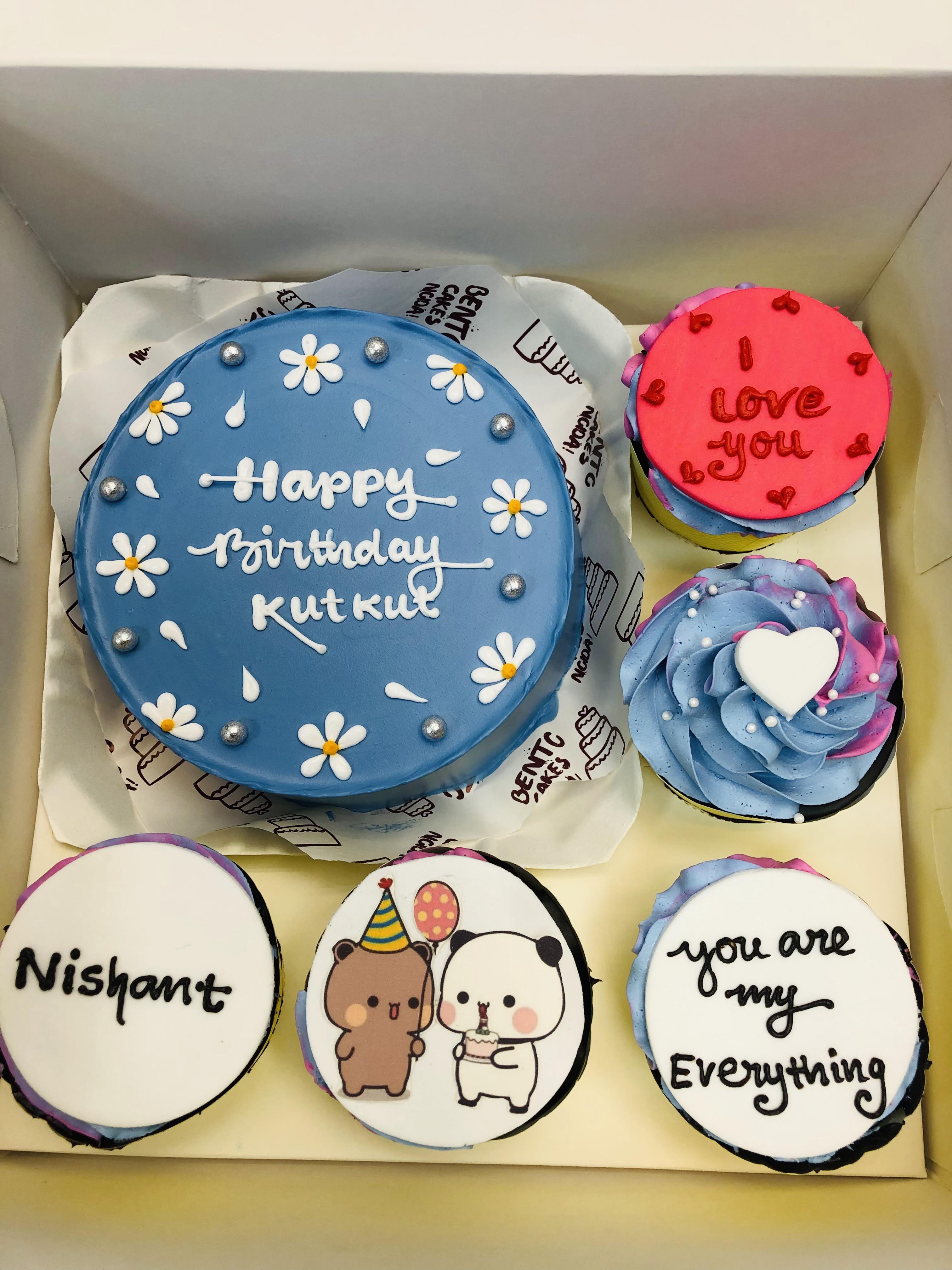 A blue frosted birthday cake adorned with white daisies, silver pearls, and the message "Happy Birthday Kutkut" in white icing, accompanied by five colorful cupcakes with unique designs and messages.