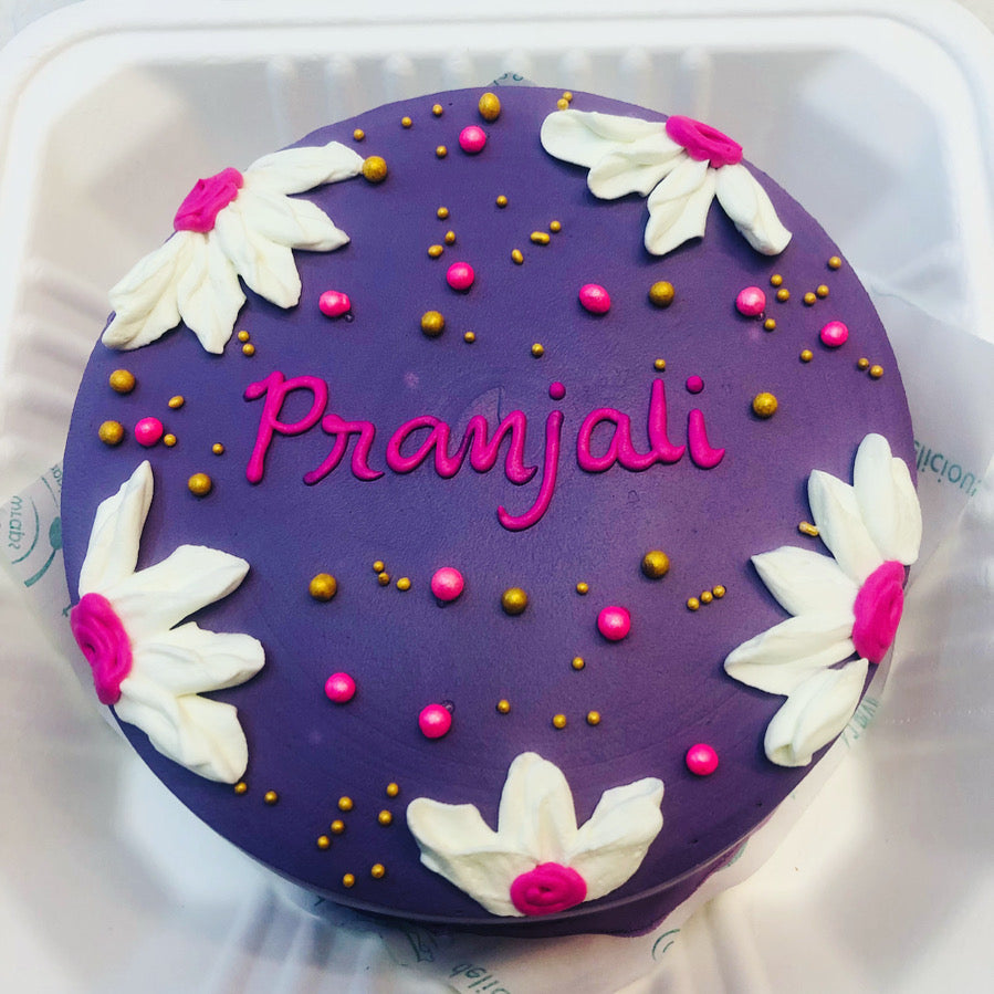 A dark purple icing cake is placed in a bento box and a white pink floral decorated on it with a "Pranjali" text written with pink icing on it.