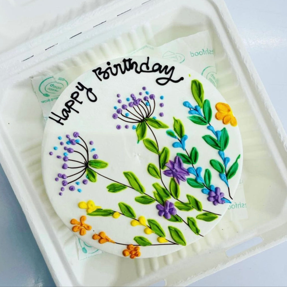 A colorful birthday cake in a bento box, adorned with vibrant flowers design on top, ready for celebration.