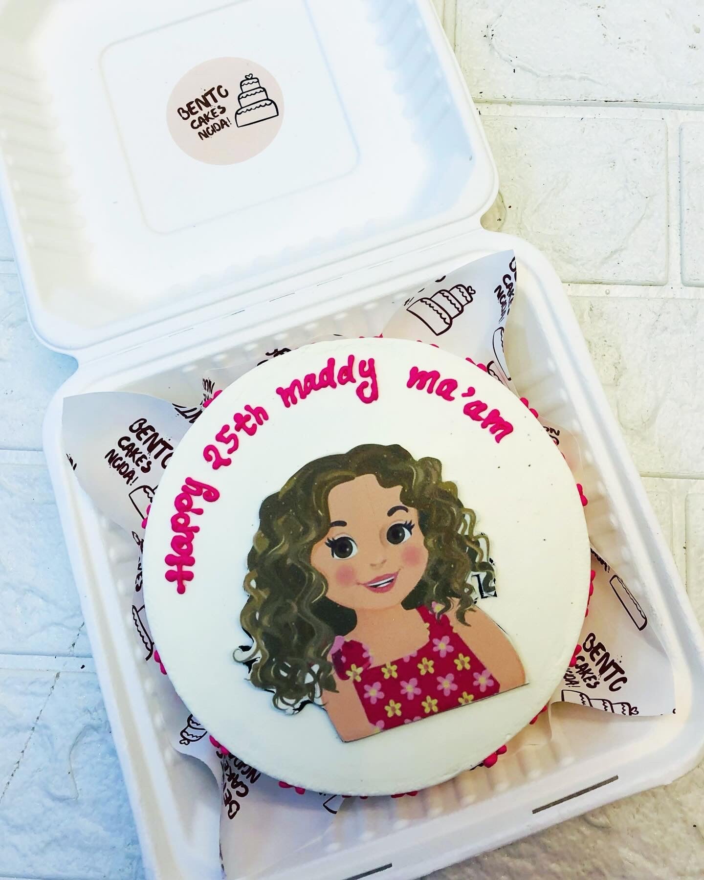 A custom birthday cake featuring a cartoon-style illustration of a girl with curly hair in a red dress with yellow flowers. The cake has pink text reading ' Happy 25th maddy ma'am' on a white icing background.