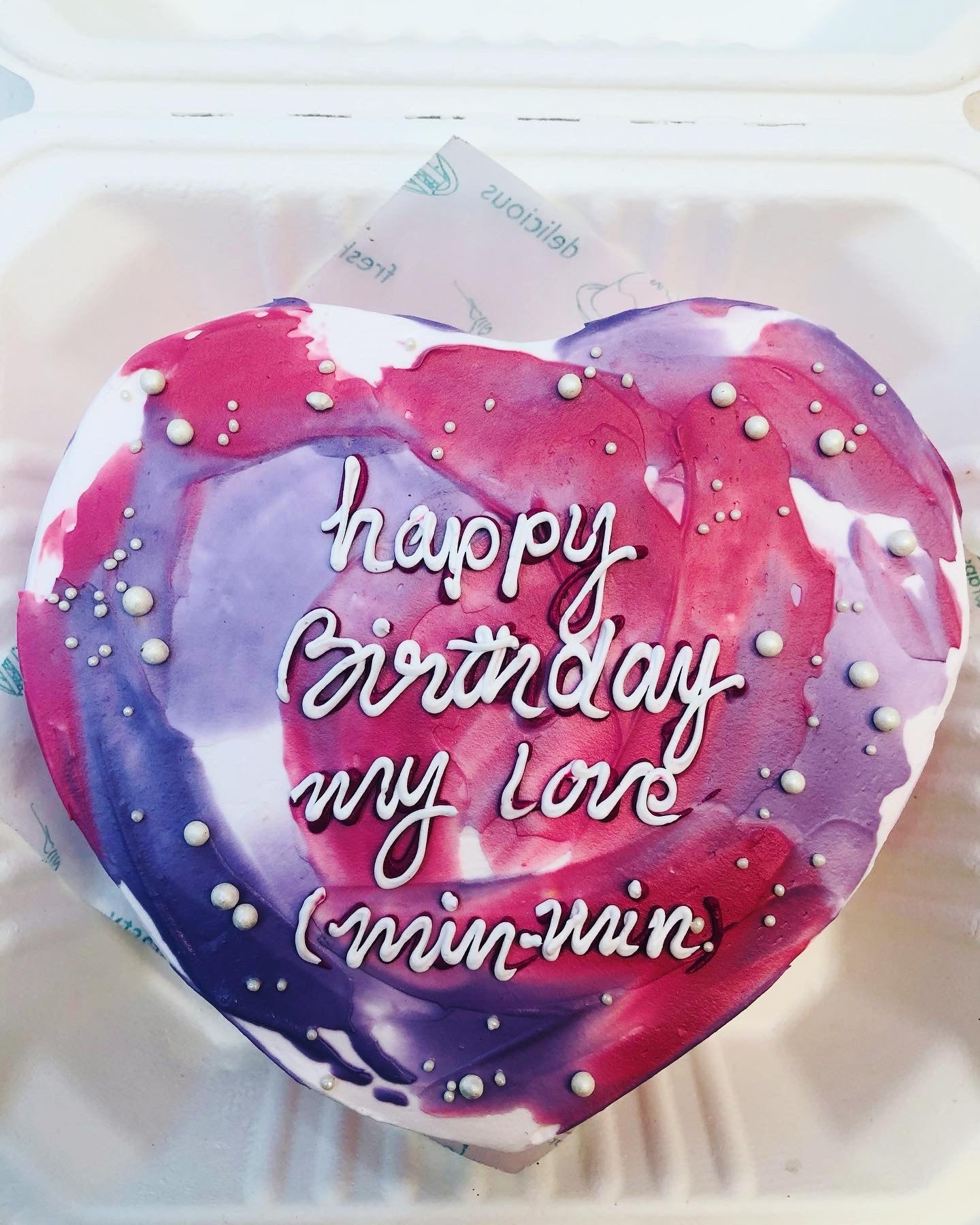 Heart-shaped birthday cake with a purple and pink marbled design, decorated with white icing text that reads "happy Birthday my love (min-min)" and small white pearls.