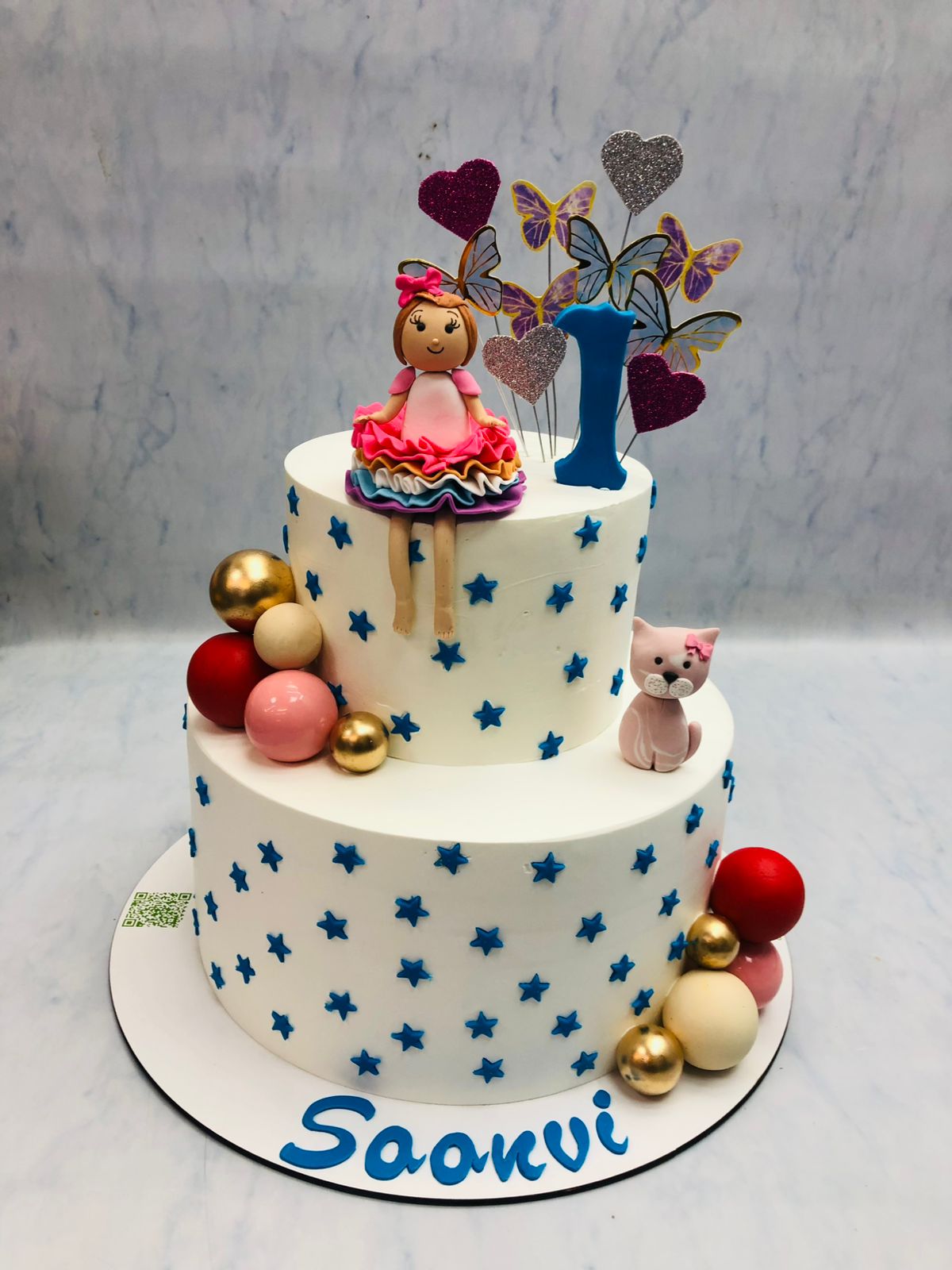 A white cake adorned with a doll on top and a cat on side, showcasing a delightful and whimsical dessert presentation.