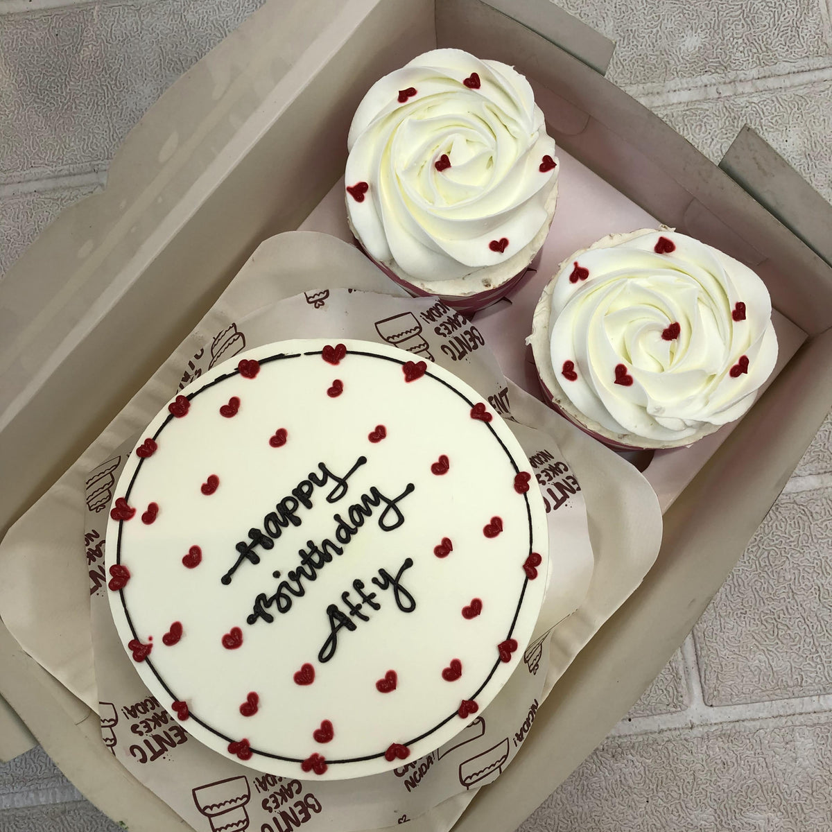 A boxed set containing a small round white cake and two cupcakes. The cake has red heart decorations and the text "Happy Birthday Affy" in black icing. The cupcakes feature white frosting with red heart decorations, all placed on decorative parchment paper with the phrases "GETTING CAKE IN THE MORNING" and "NEON."