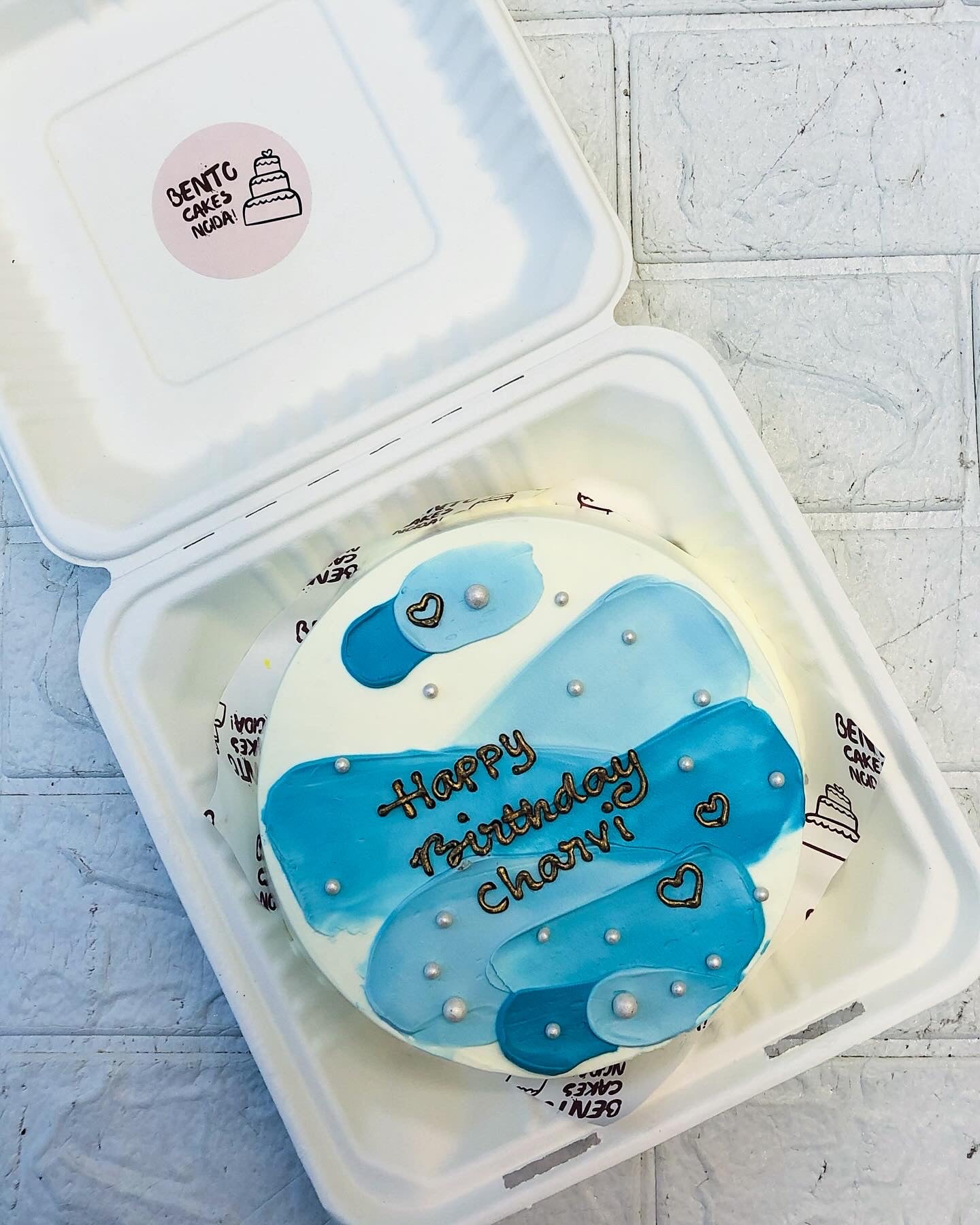 A beautifully decorated round birthday cake featuring blue and white abstract designs, placed inside a white bento box. The cake has 'Happy Birthday Charvi!' written in brown icing, with delicate pearl-like decorations and heart shapes.