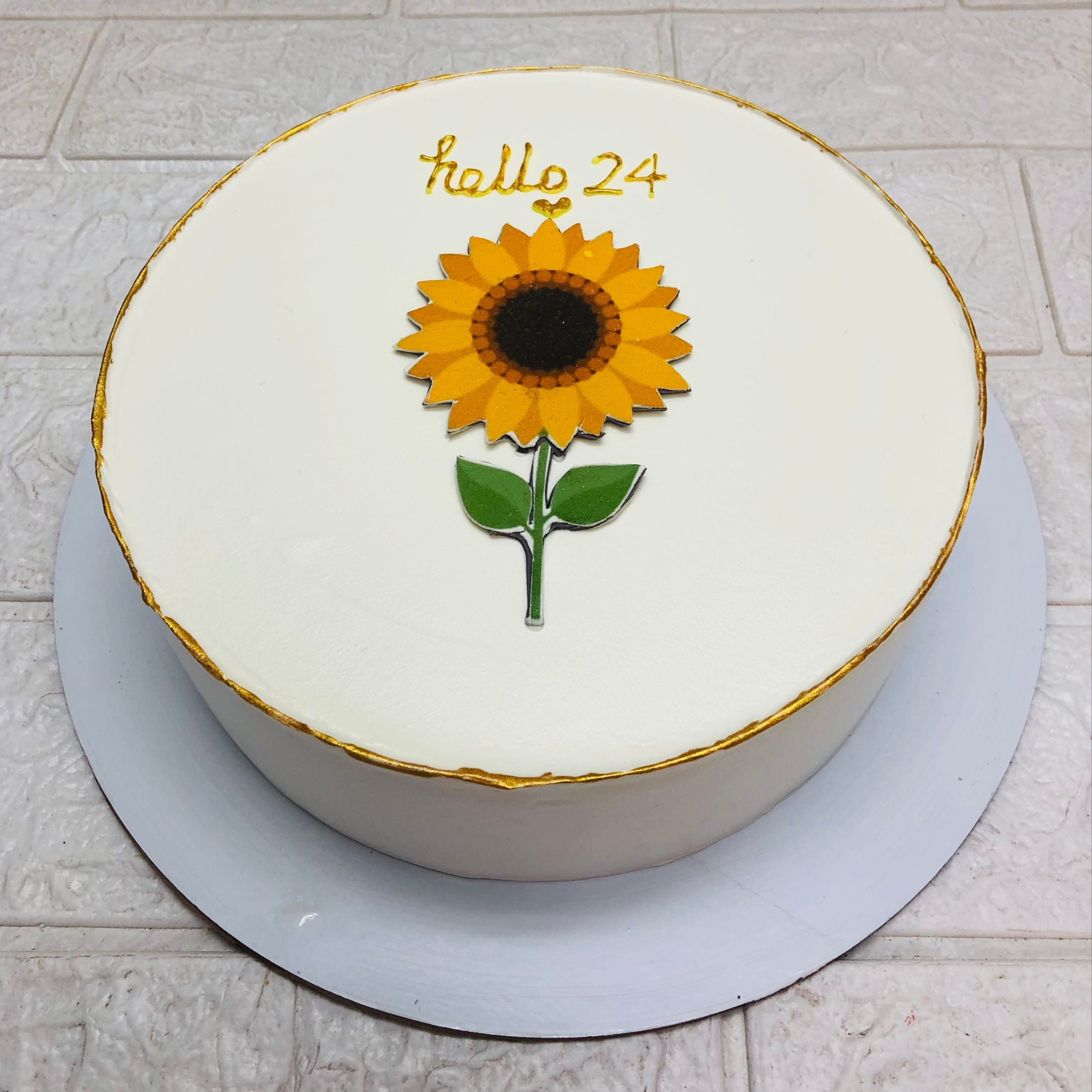 A round white cake designed with a sunflower sticker on the center of cake and a "hello 24" text written with golden icing and cake itself surrounded by golden line.