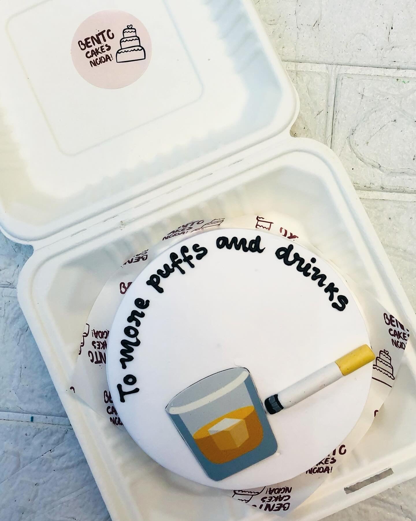A white bento cake is designed with a cigeetgre and alcohol glass printb with an unique text.