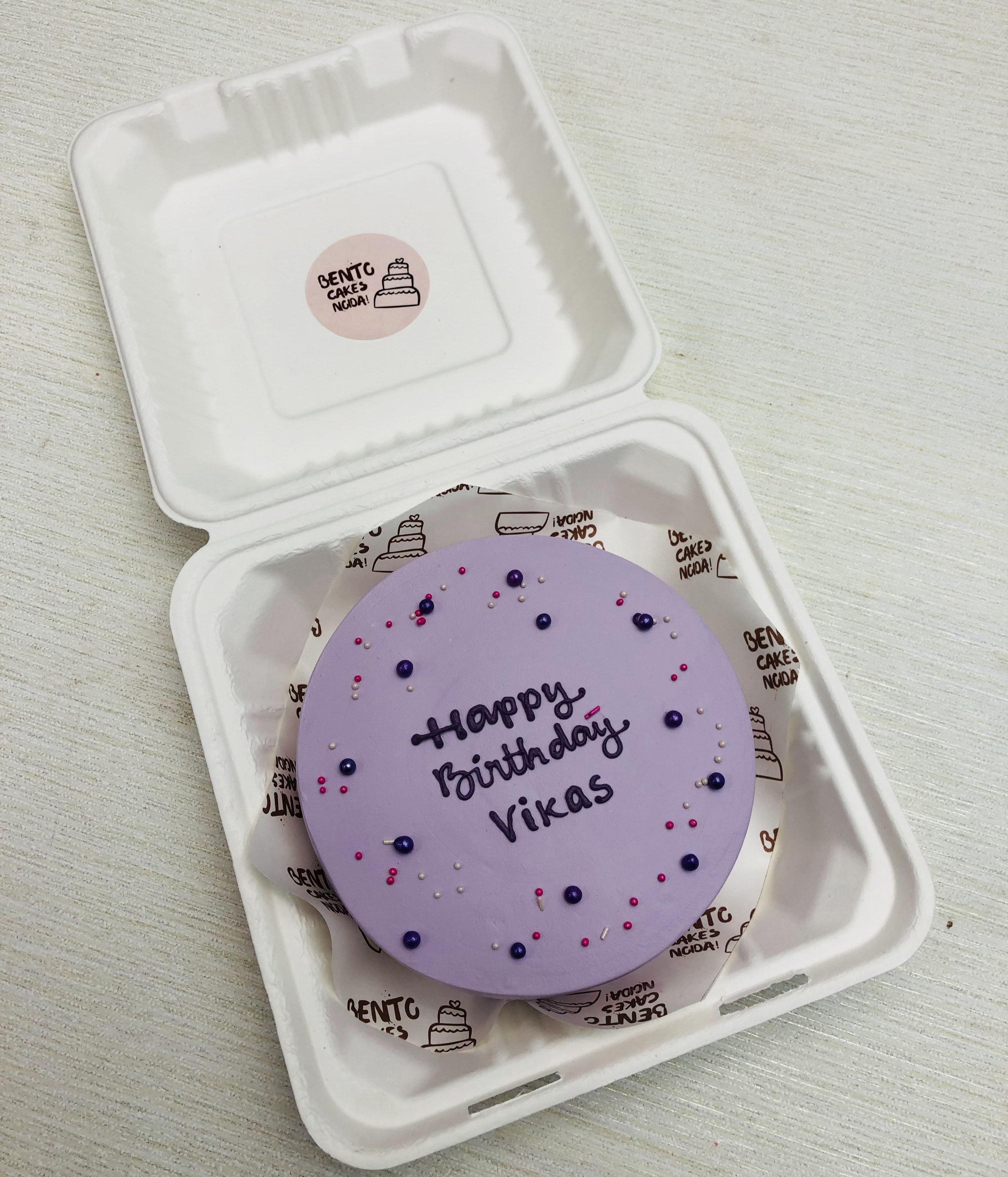 A light purple bento cake with purple text of "Happy Birthday Vikas" and decorated with purple and pink spinkles.