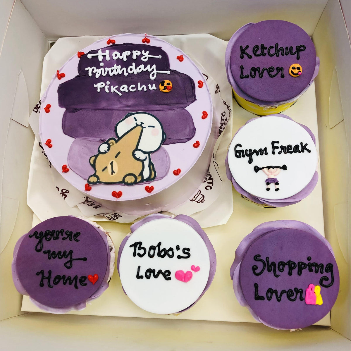 A Purple Birthday Bento Combo Cake set featuring one cake and five cakes. The large cake has purple frosting with the text "Happy Birthday Pikachu" in white icing and an illustration of a character holding a piece of cheese surrounded by red hearts. The smaller cakes are decorated in purple and white themes, each with unique text and designs, such as "Ketchup Lover," "Gym Freak," and others.