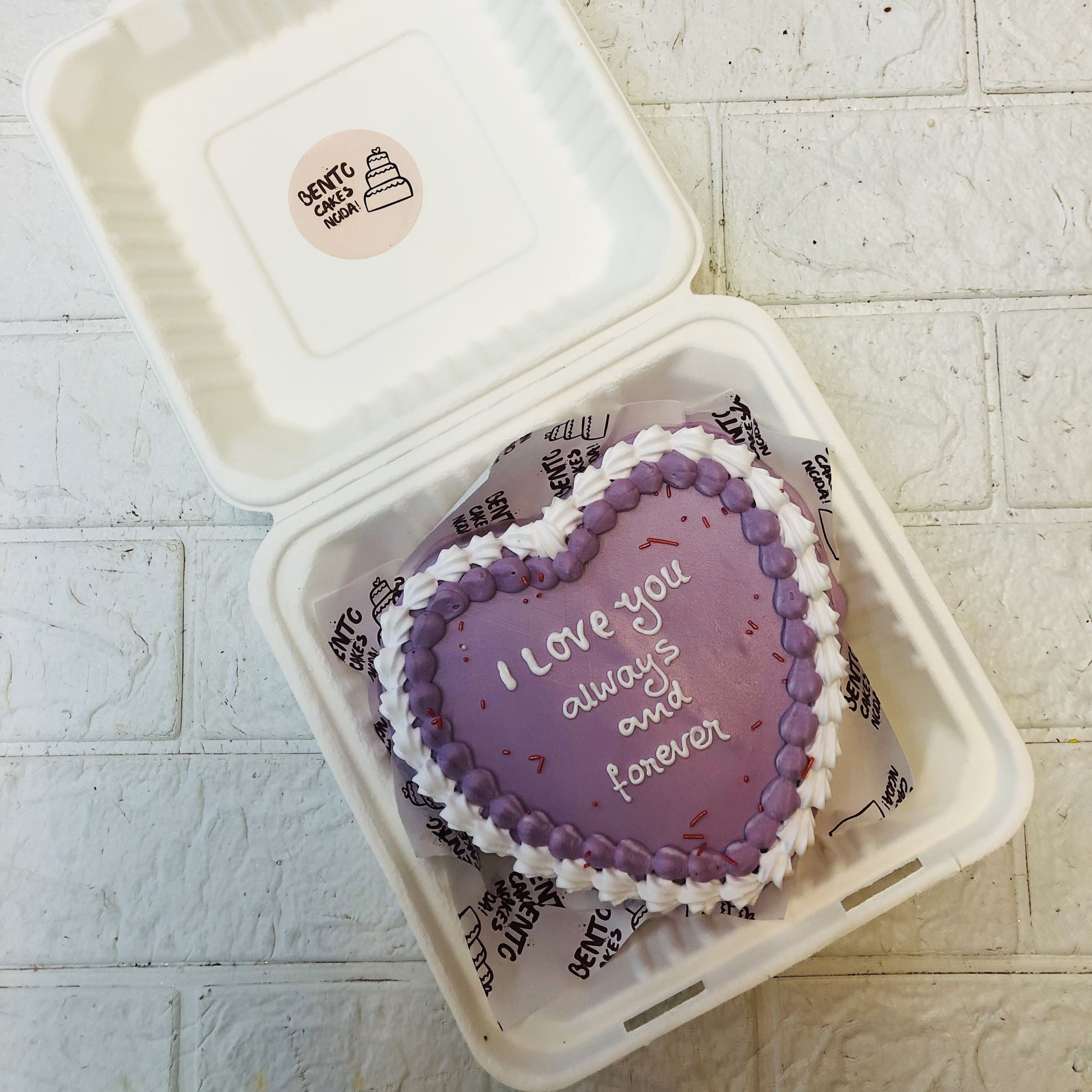 A purple heart shape cake with purple and white boundry and a loving text written on it.