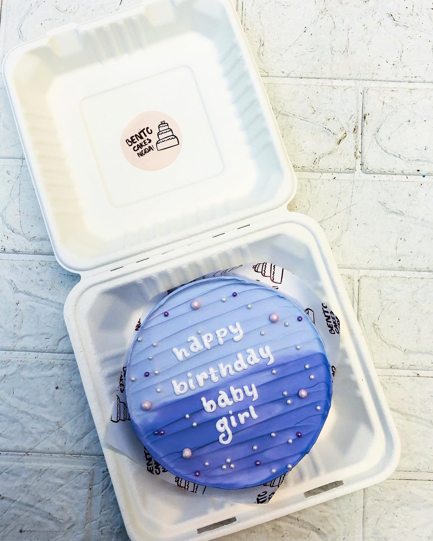A round purple vegan birthday cake with 'happy birthday baby girl' written in white icing, placed inside a white takeout box. The cake is decorated with small white and purple dots.