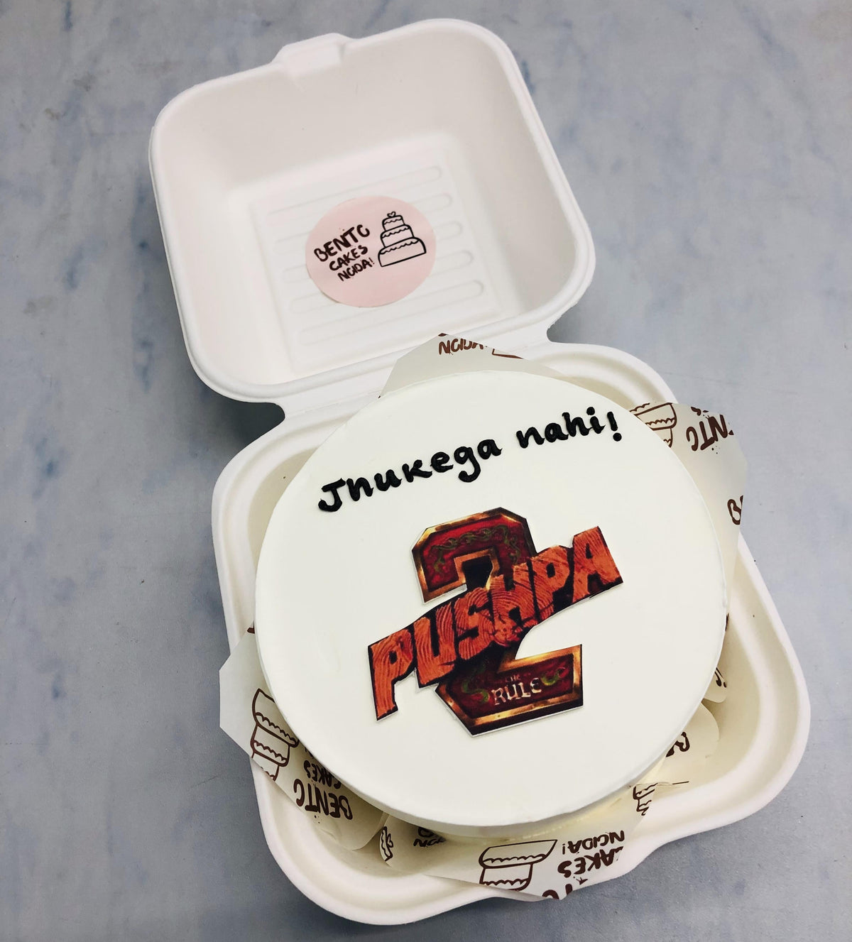 A white bento cake is designed on a movie theme "Pushpa 2" with its trending dialogue.