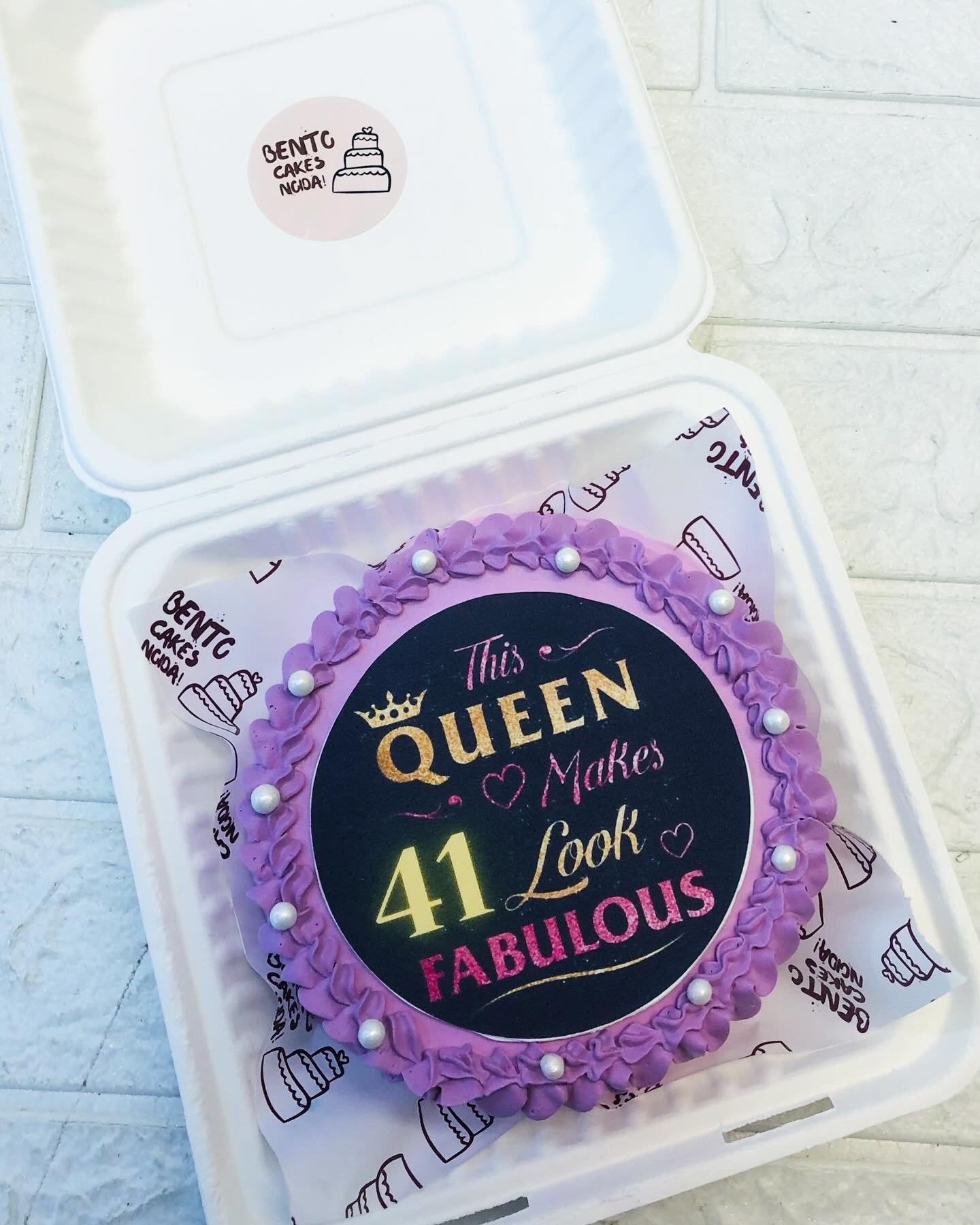 A purple birthday cake with the text 'This Queen Makes 41 Look Fabulous' written on it, decorated with pearl-like beads and placed in a white bento box.