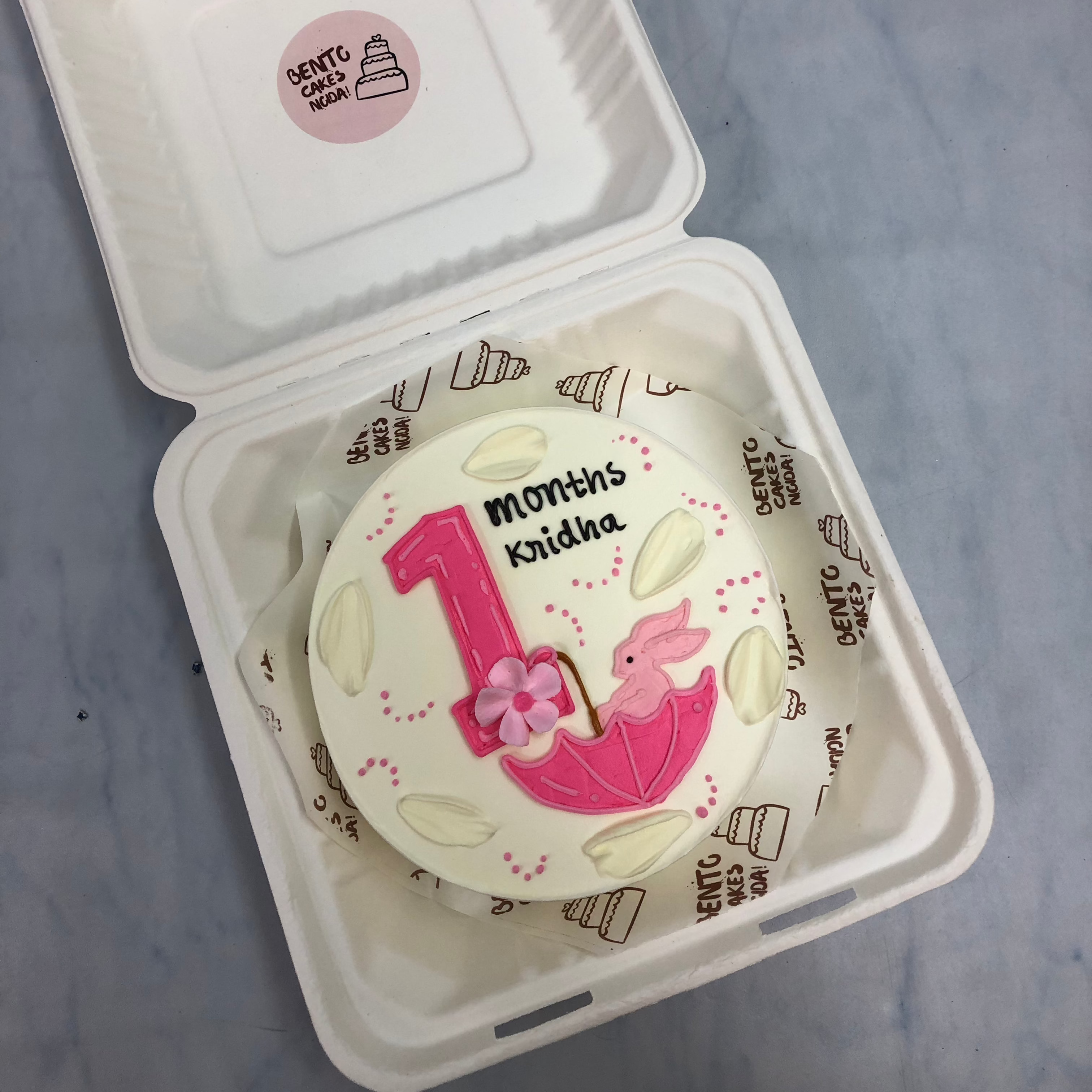 An offehite cake with a pink rabbt in pink umbrella and is designed for 1 month baby girl.