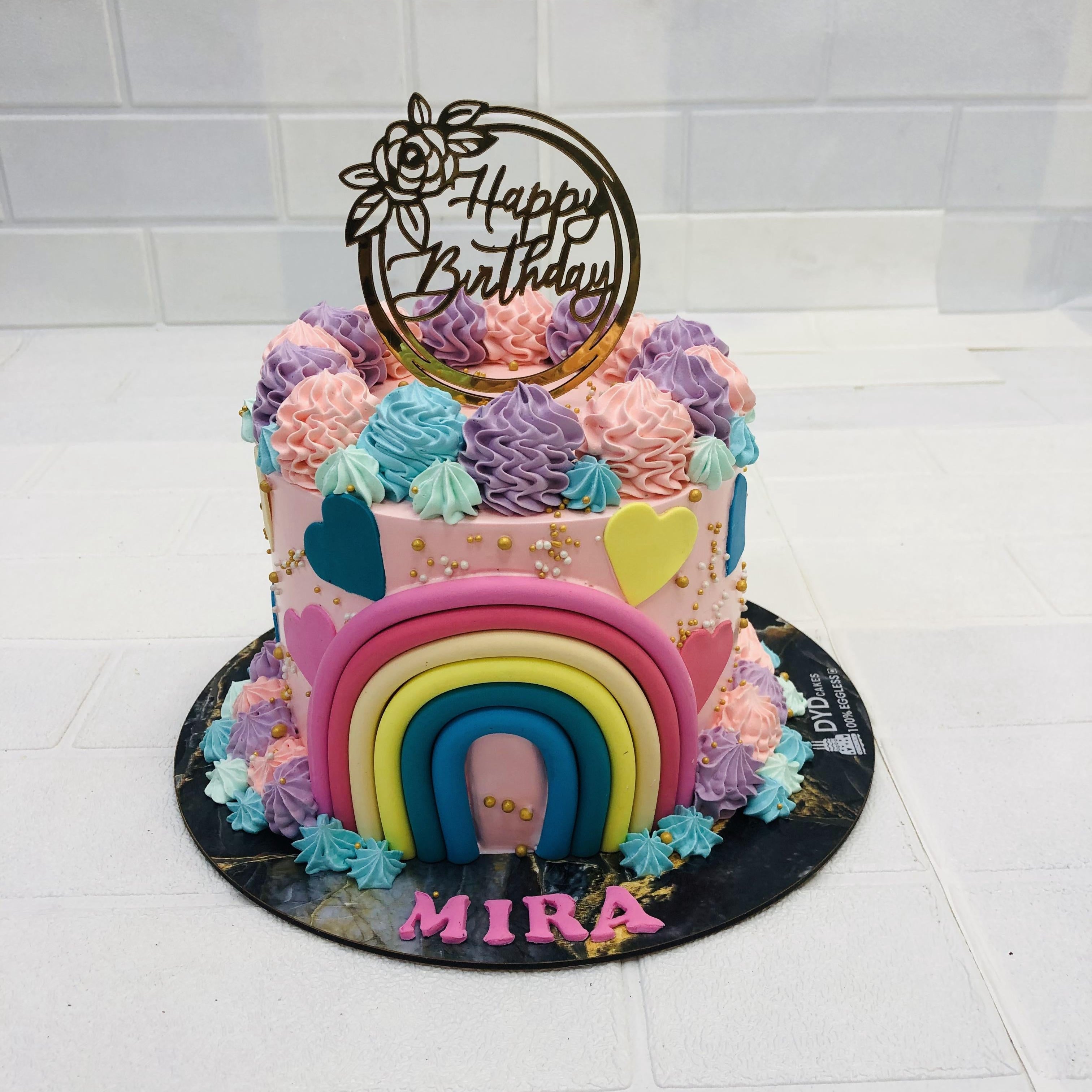 A beautifully decorated birthday cake featuring a rainbow design and pastel-colored frosting. The cake has a 'Happy Birthday' topper and the name 'Mira' written at the base."