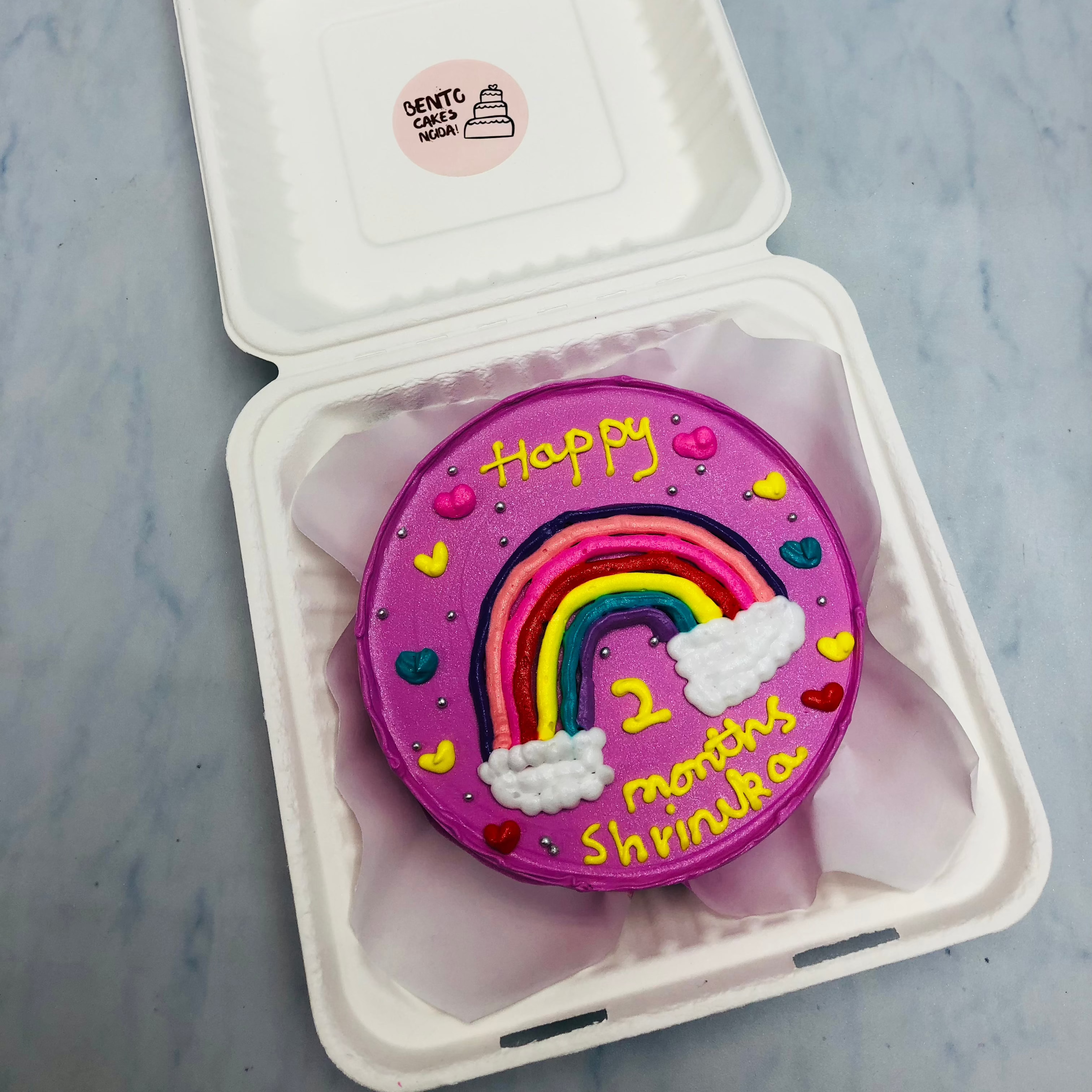 A purple bento cake with colorful rainbo and a text written with yellow icing.